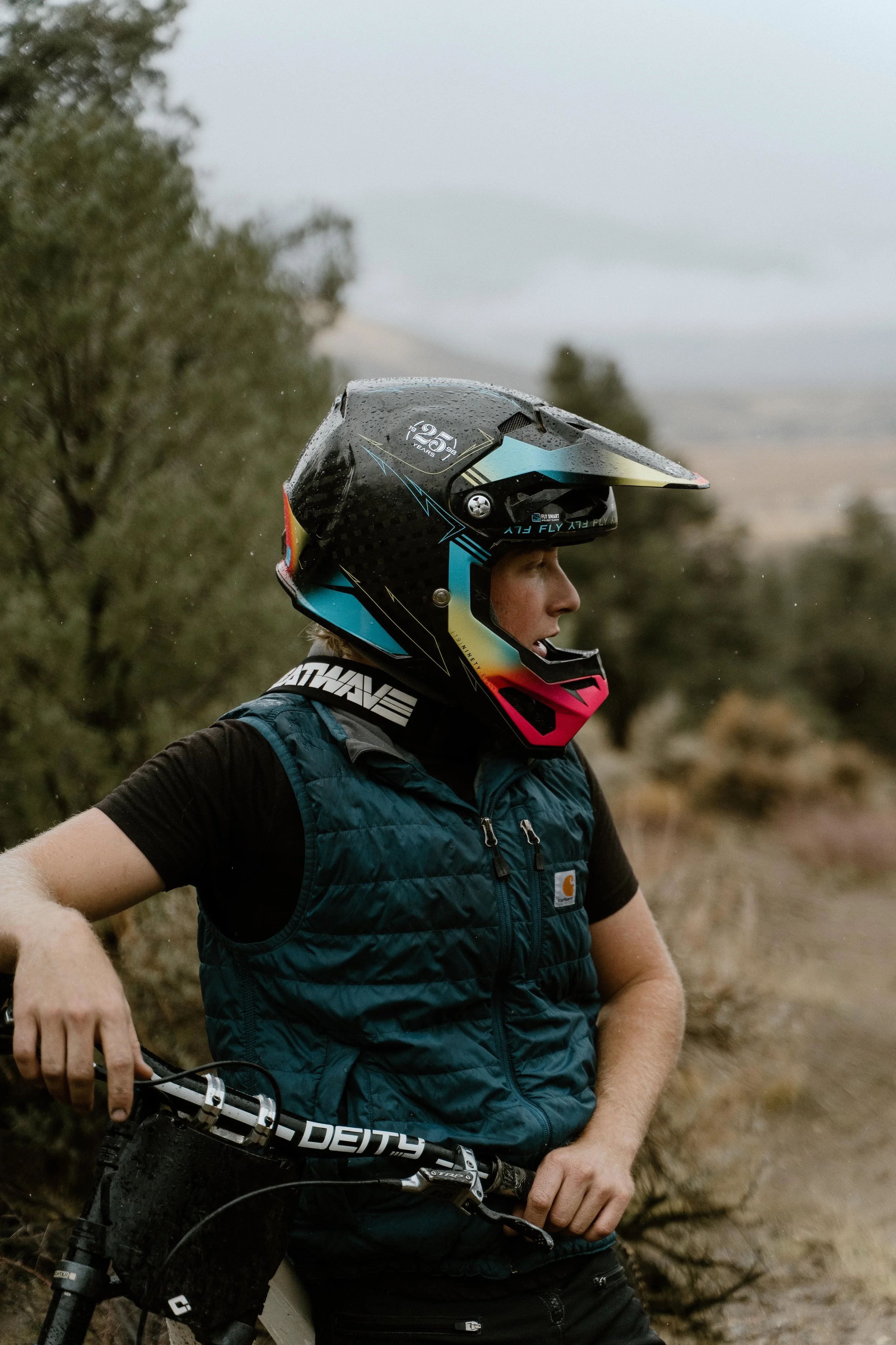 A person wearing a black and colorful full-face helmet, black t-shirt, and a blue vest is standing outdoors with a mountain bike, in a natural, slightly rainy environment.