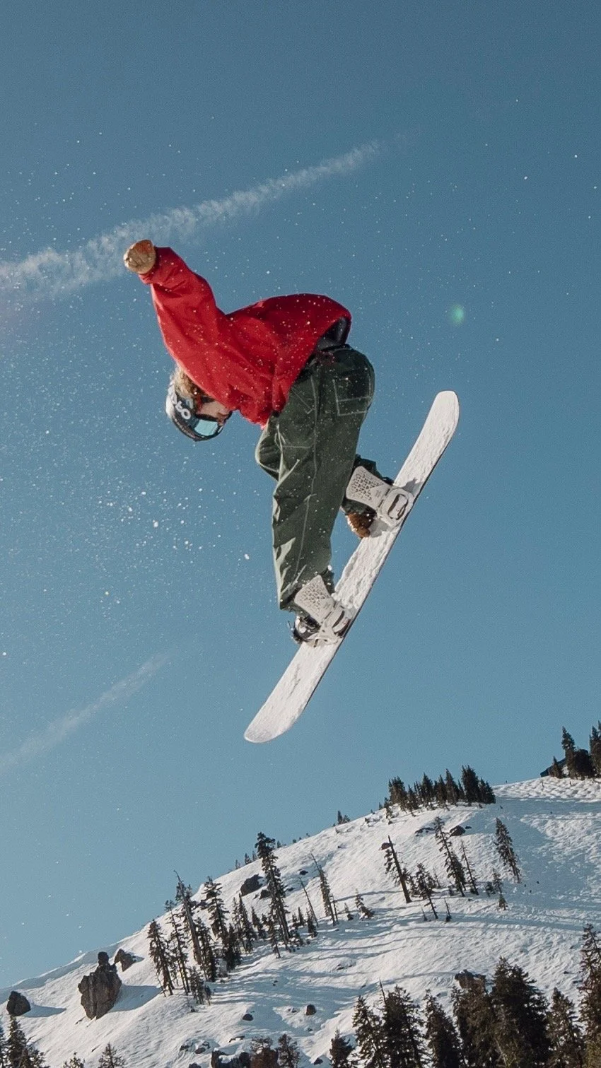 A snowboarder wearing a red jacket and black pants jumping in mid-air against a snowy mountain landscape and clear blue sky.