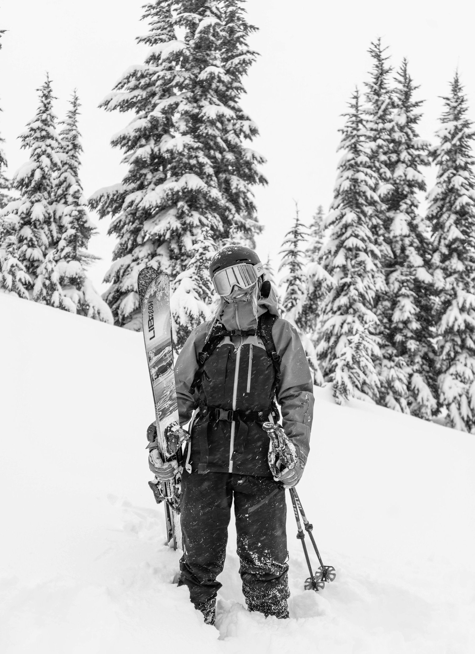 A person wearing ski gear, including a helmet, goggles, and a winter jacket, stands in snow holding ski poles and a pair of skis, with snow-covered trees in the background.