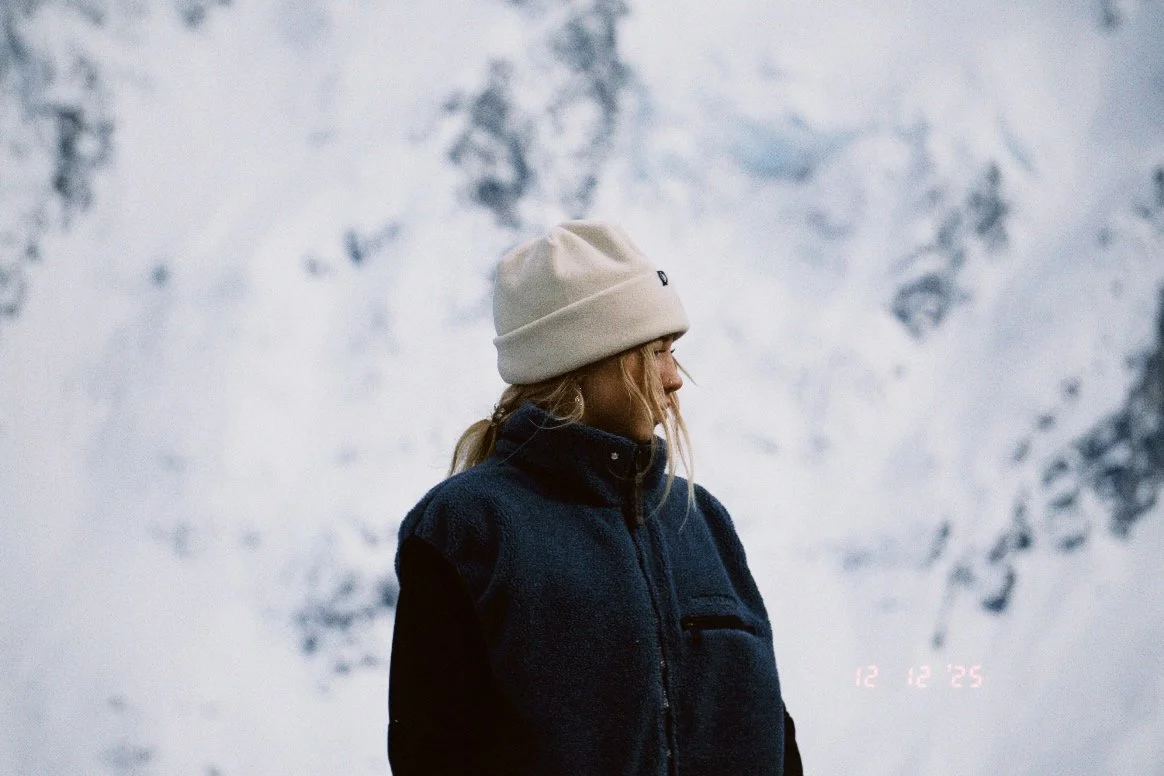A woman with blonde hair wearing a beige beanie and a navy fleece vest, standing outdoors against a snow-covered mountain background.