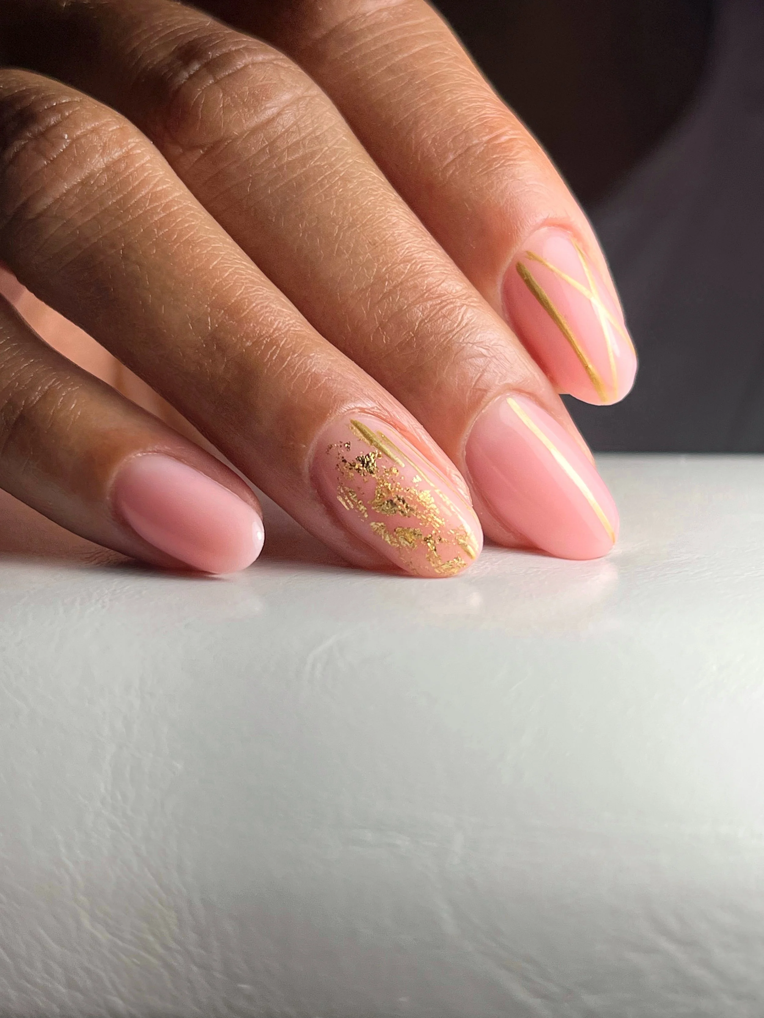 Close-up of a person's hand with a manicured display of nails featuring a light pink base, some with gold accents and decorative gold foil on the ring finger.