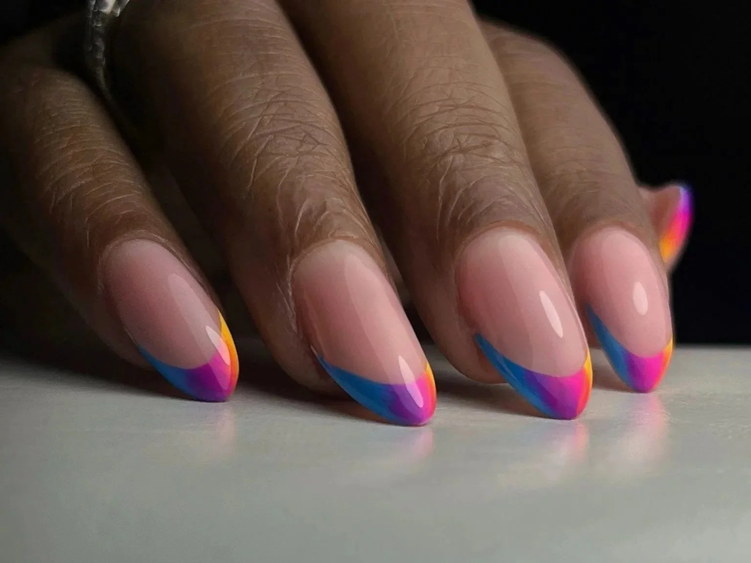 Close-up of a hand with nails painted in a French manicure style with colorful rainbow tips, resting on a light surface.