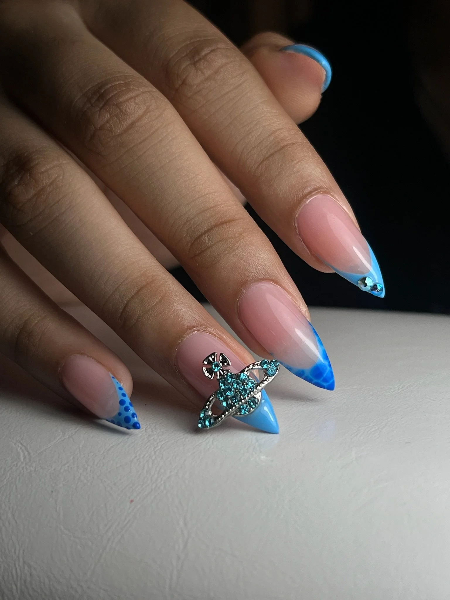 Fingernails with blue and white gradient acrylic nails, featuring pointed tips, adorned with a silver and turquoise space-themed ring. The skin is medium-toned with visible creases on the fingers.