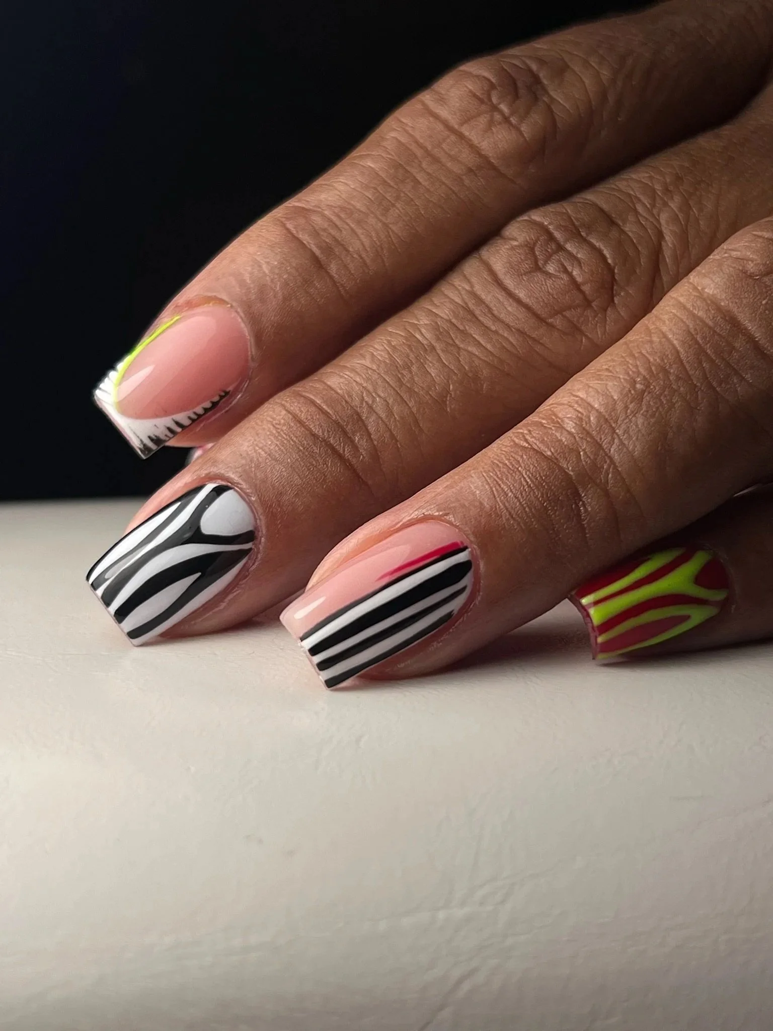 Close-up of a hand showcasing multi-colored, patterned nail art on short, square-shaped nails. The designs include black and white zebra stripes, pink and black lines, and yellow and green patterns.
