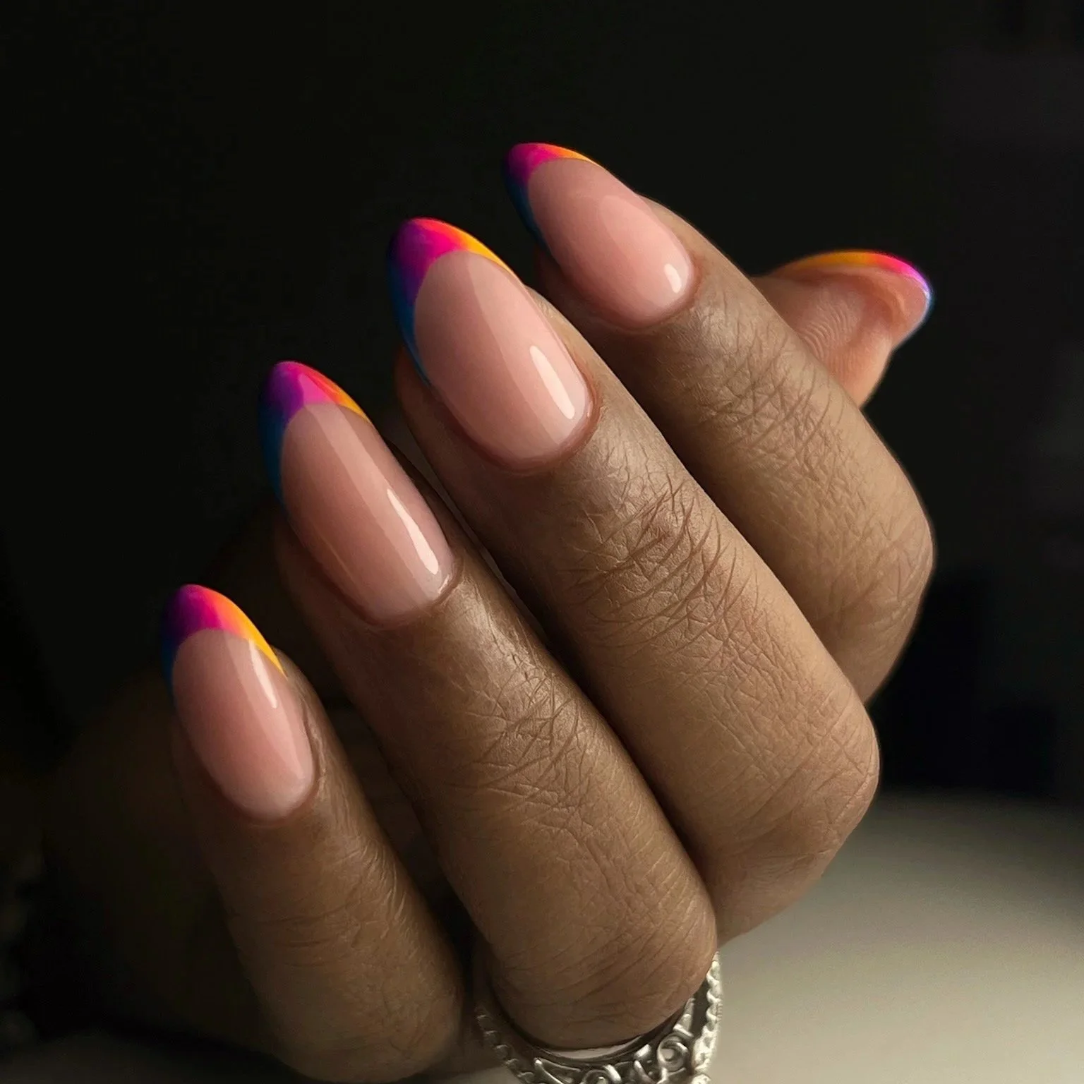 Close-up of a hand with almond-shaped nails painted with gradient pink, orange, yellow, and blue tips.