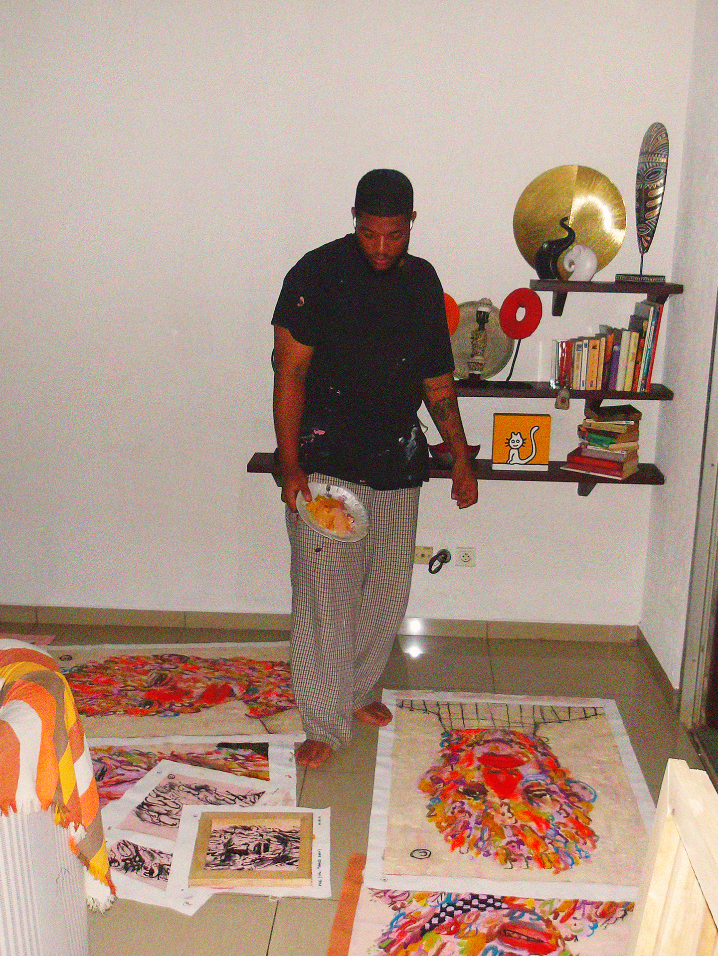 A man standing on a beige tiled floor in a room, surrounded by colorful artwork displayed on the floor and a wall shelf with decorative items and books.