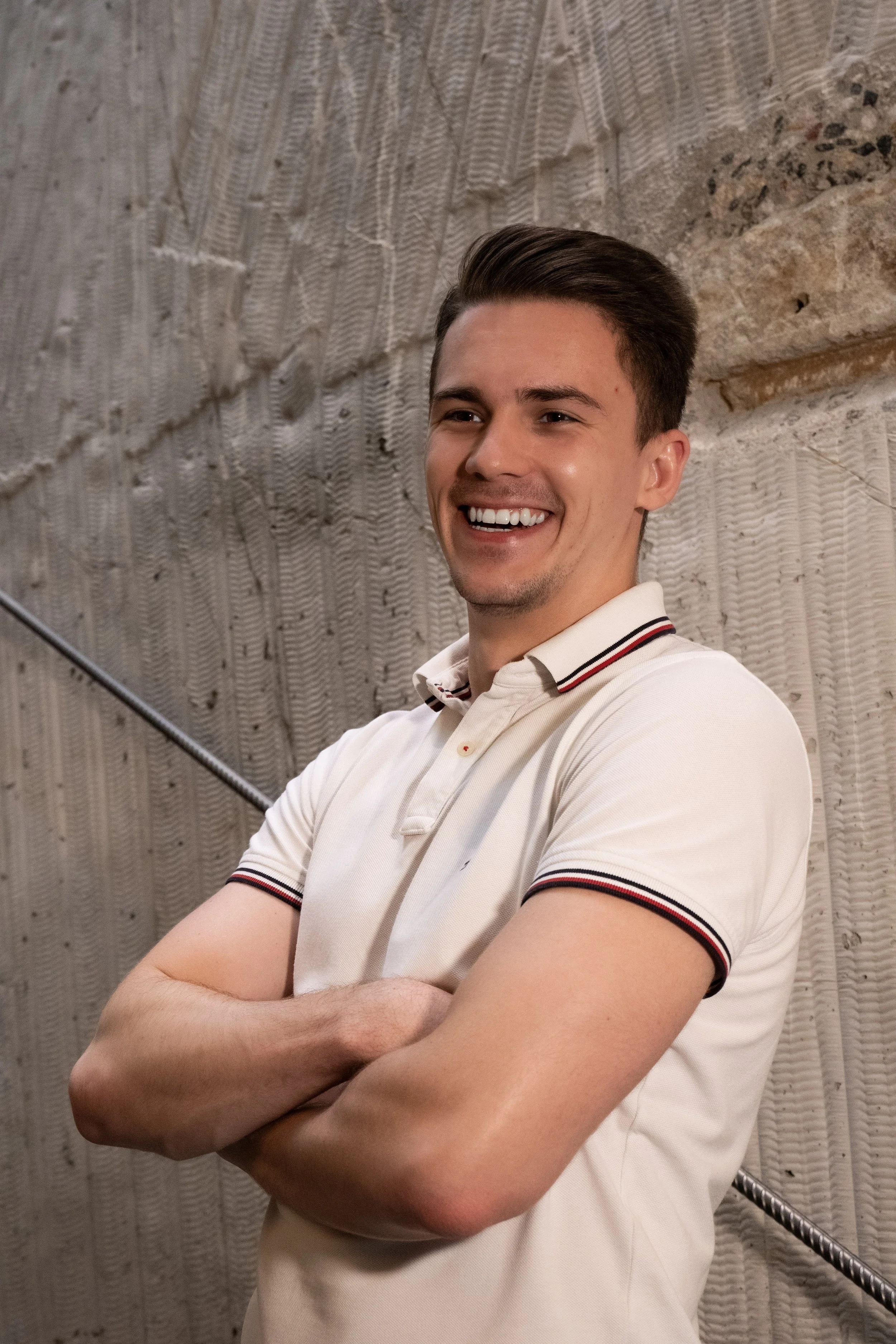 Smiling young man with arms crossed, wearing a white polo shirt with red and black trim, standing against a textured stone wall.