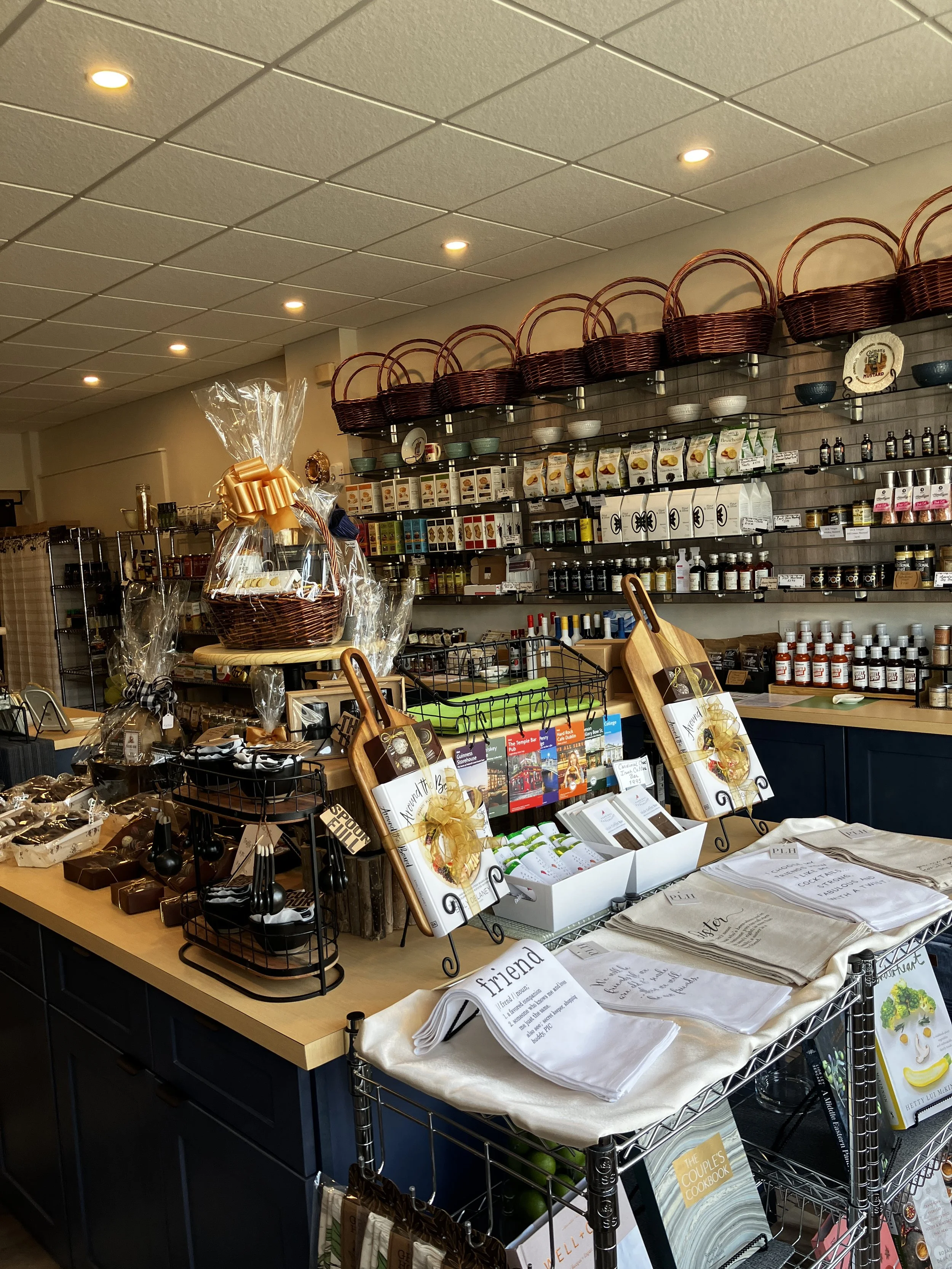 Interior display at The Uncommon Pantry showing hand-curated gift baskets wrapped in cellophane with gold bows, surrounded by shelves of gourmet spices, sauces, teas, cookbooks, and kitchen towels.