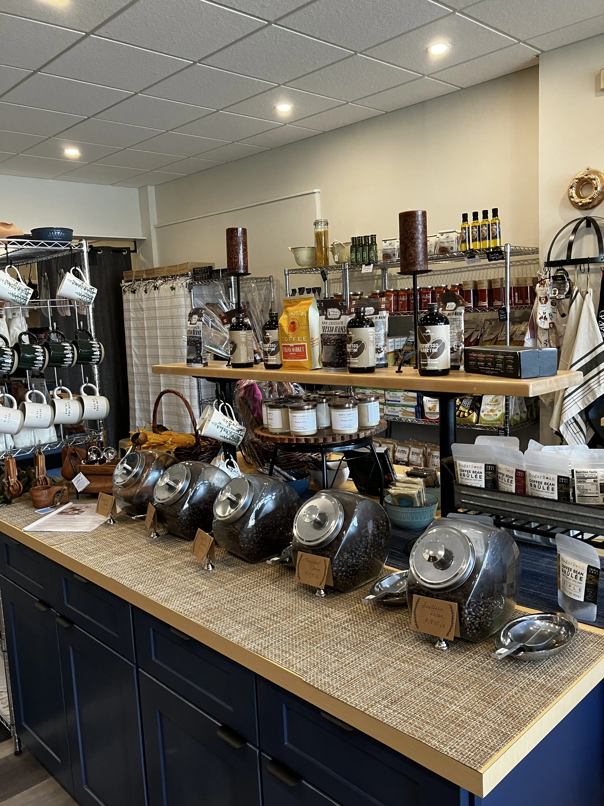 Coffee and confection display at The Uncommon Pantry featuring large jars of whole bean coffees like Espresso and Brûlée, packaged beans, chocolates, mugs, and specialty items on tiered wooden shelves.