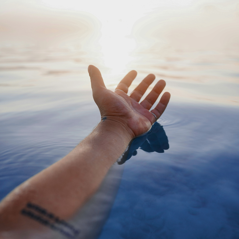 A person's hand reaching out over water with a bright sun or light source in the background.