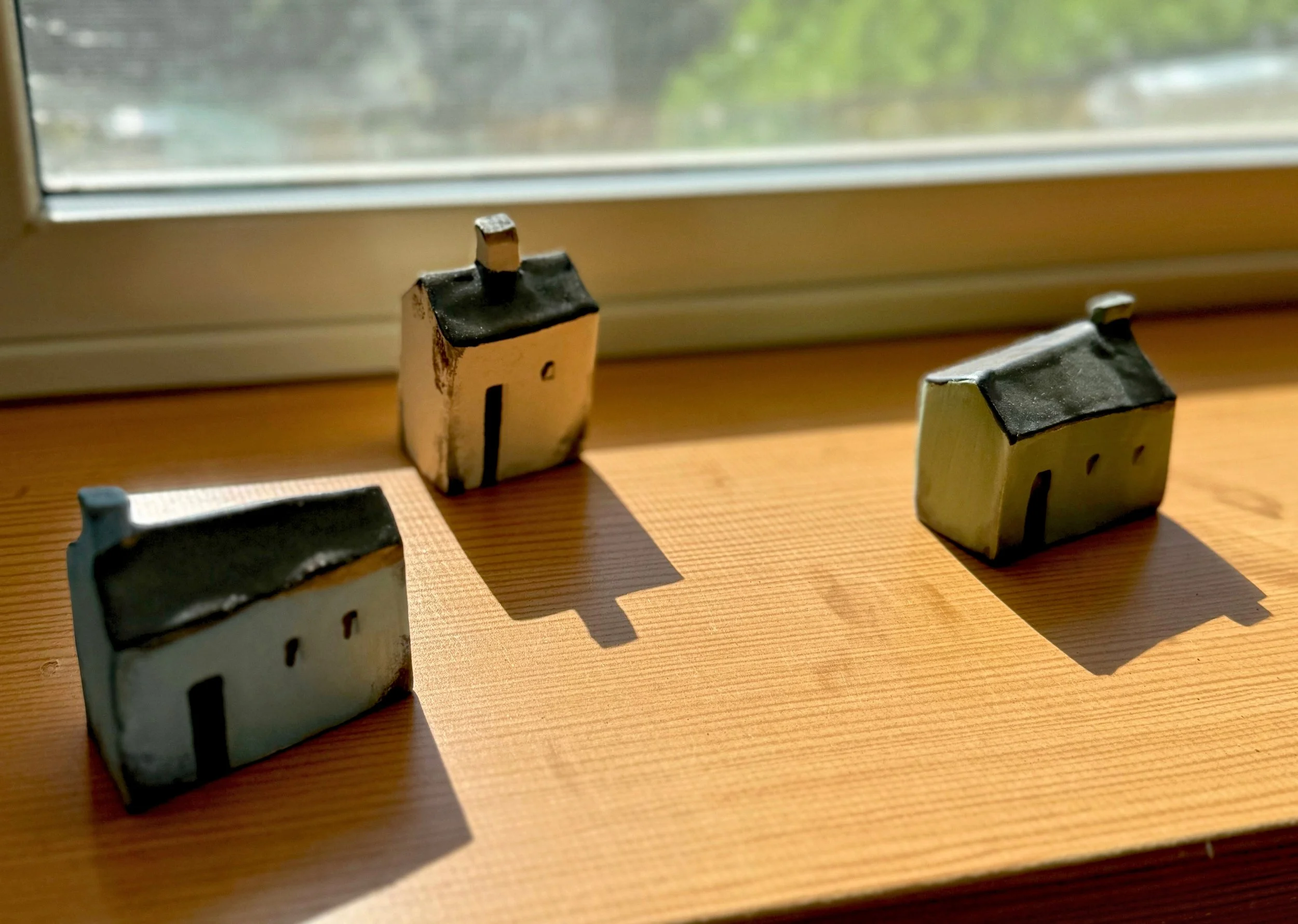 Three small ceramic house figurines placed on a wooden windowsill with sunlight casting shadows, viewed from an angle. The houses are in different colors and shapes, with the window in the background.