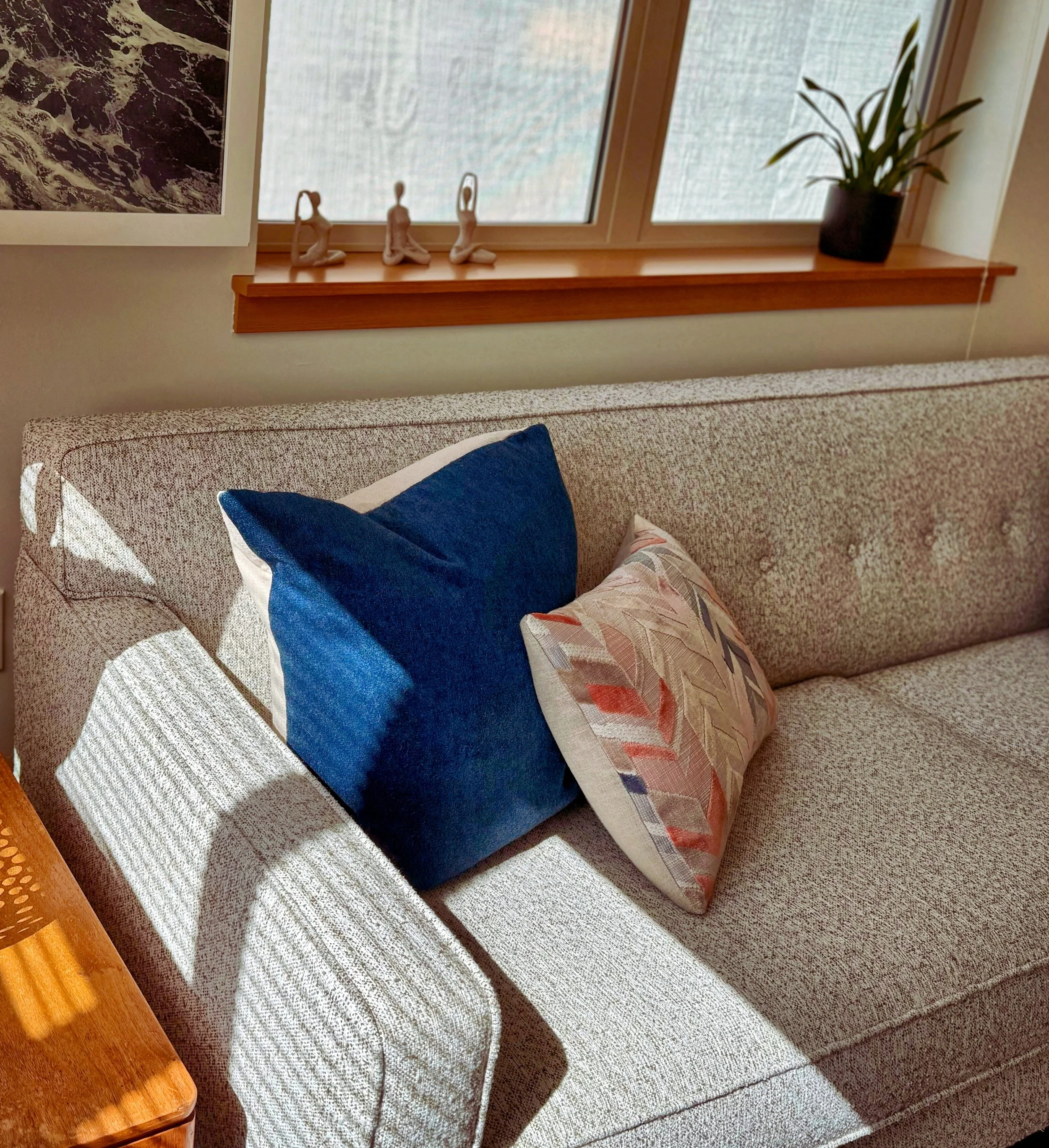 A beige fabric sofa with two decorative pillows, one navy blue and one with a multicolored leaf pattern. Sunlight creates shadows on the sofa. A wooden table with an orange hue is partially visible in the foreground. Behind the sofa, a window with fr