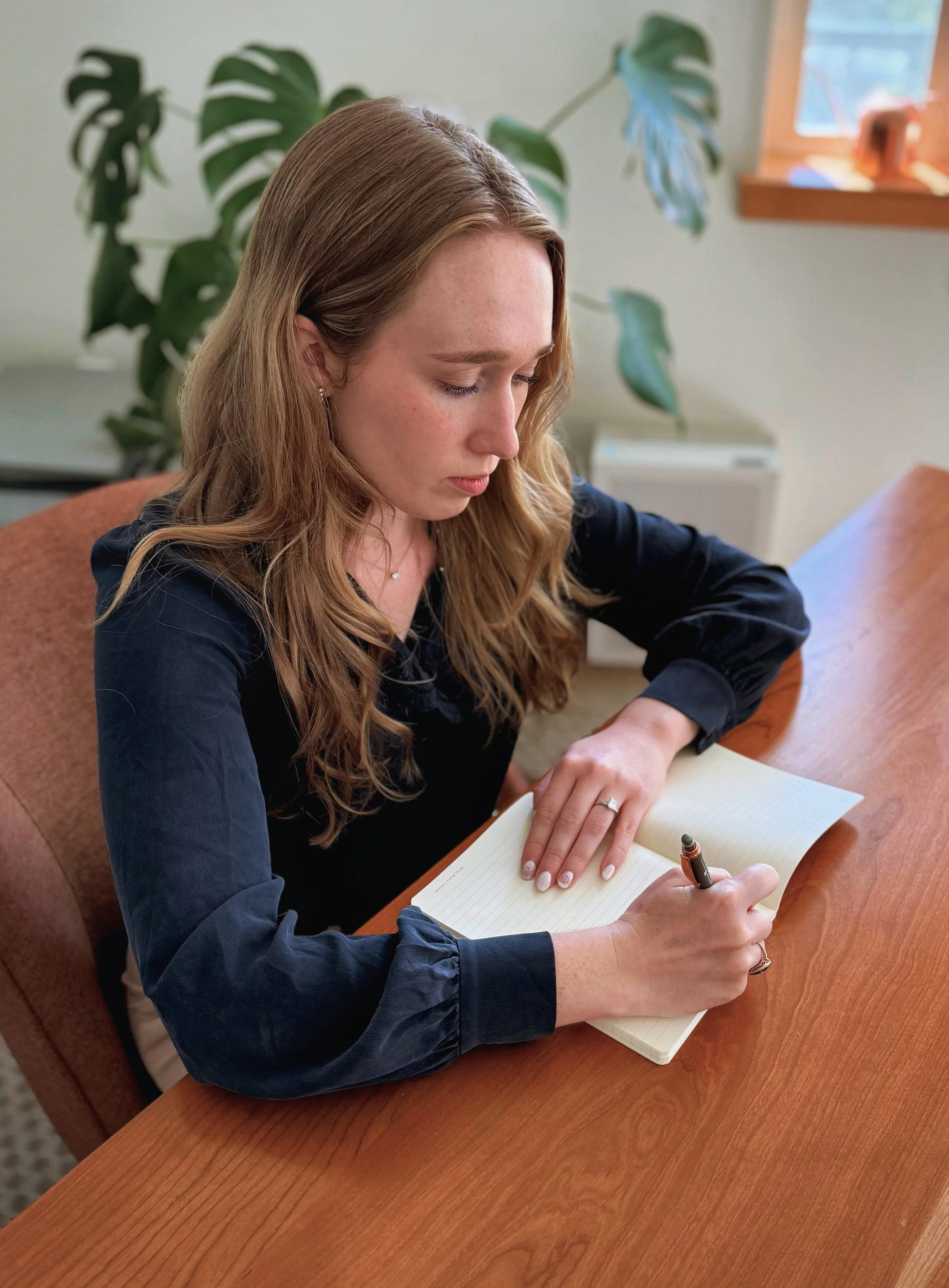 A woman with long wavy hair and a ring on her left hand is writing in a lined notebook with a black pen at a wooden table. She is wearing a black blouse with puffed sleeves. Behind her, there are large green houseplants and a window with wooden trim.