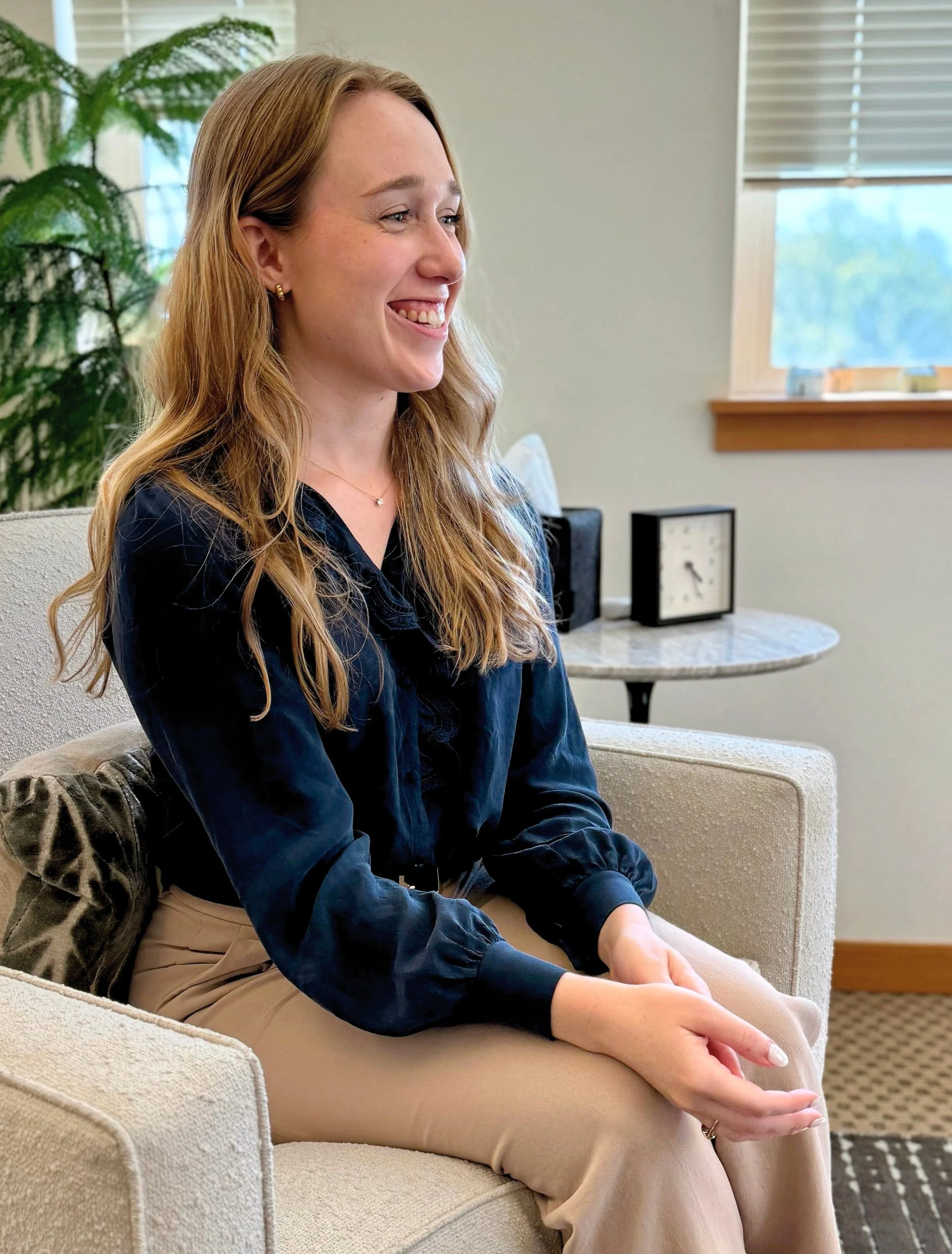 A woman with long, wavy red hair sitting on a beige armchair, smiling and wearing a navy blue blouse and tan pants, in a room with green plants and a window with blinds.