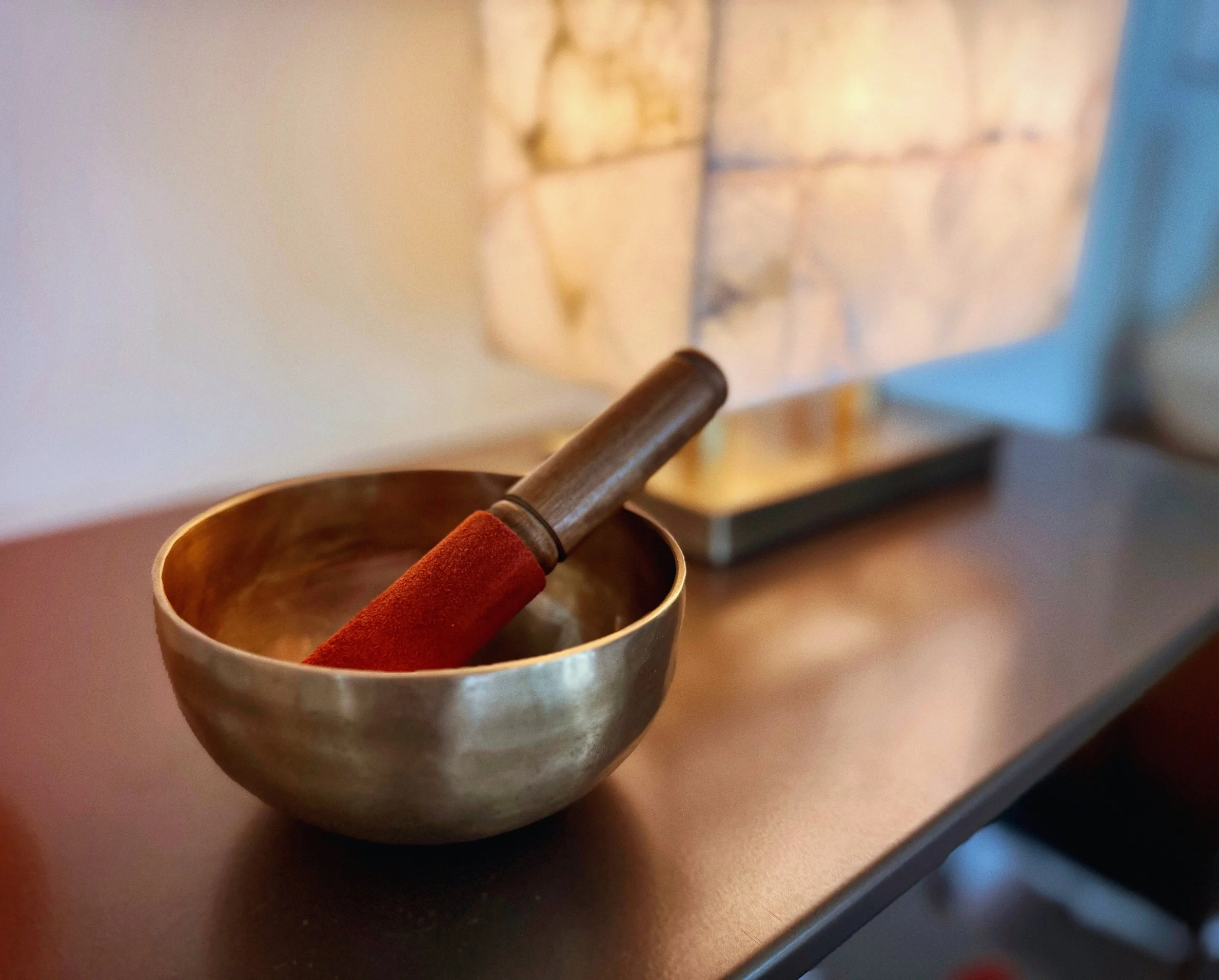 A brass singing bowl with a wooden mallet resting inside it on a wooden surface, with a marble lamp in the background.