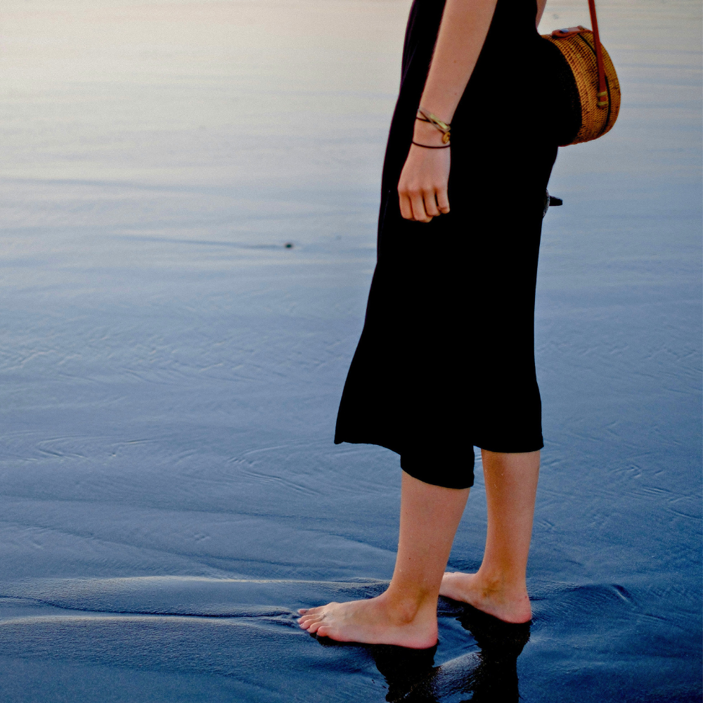 Person standing barefoot on wet sand near the water's edge, wearing a black dress and carrying a woven bag.