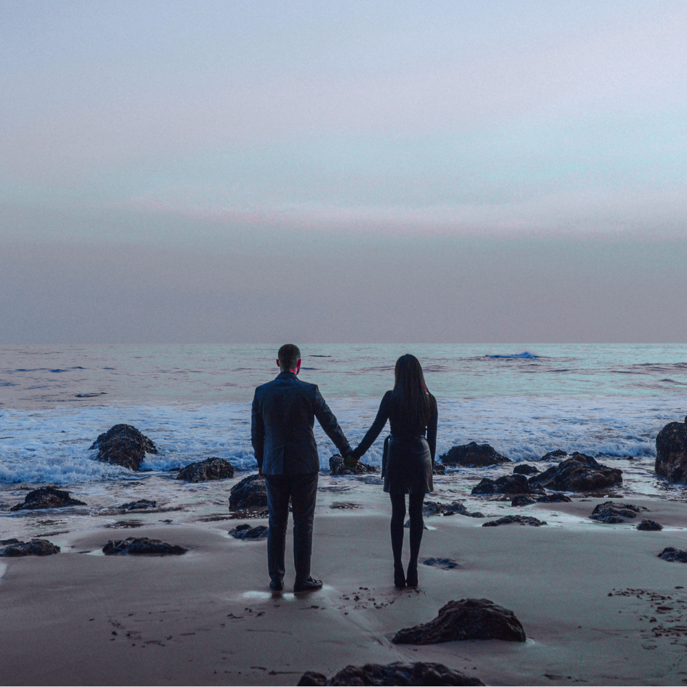 A man and woman holding hands on a beach at sunset with rocks and ocean waves.