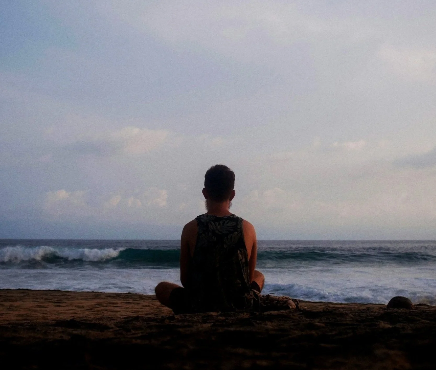 Person sitting on the beach facing the ocean, with waves and cloudy sky in the background.