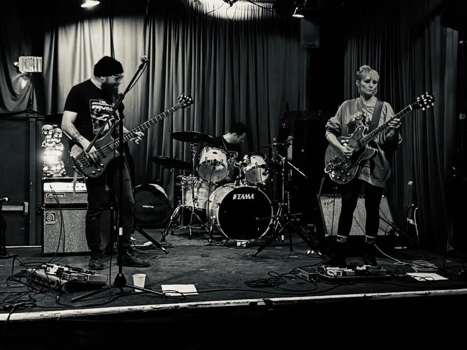 A band performing live on stage in a dimly lit venue, featuring a bassist, drummer, and guitarist. My Mistress at Valhalla Chace Levi Lacey Kay Levi Martin leal
