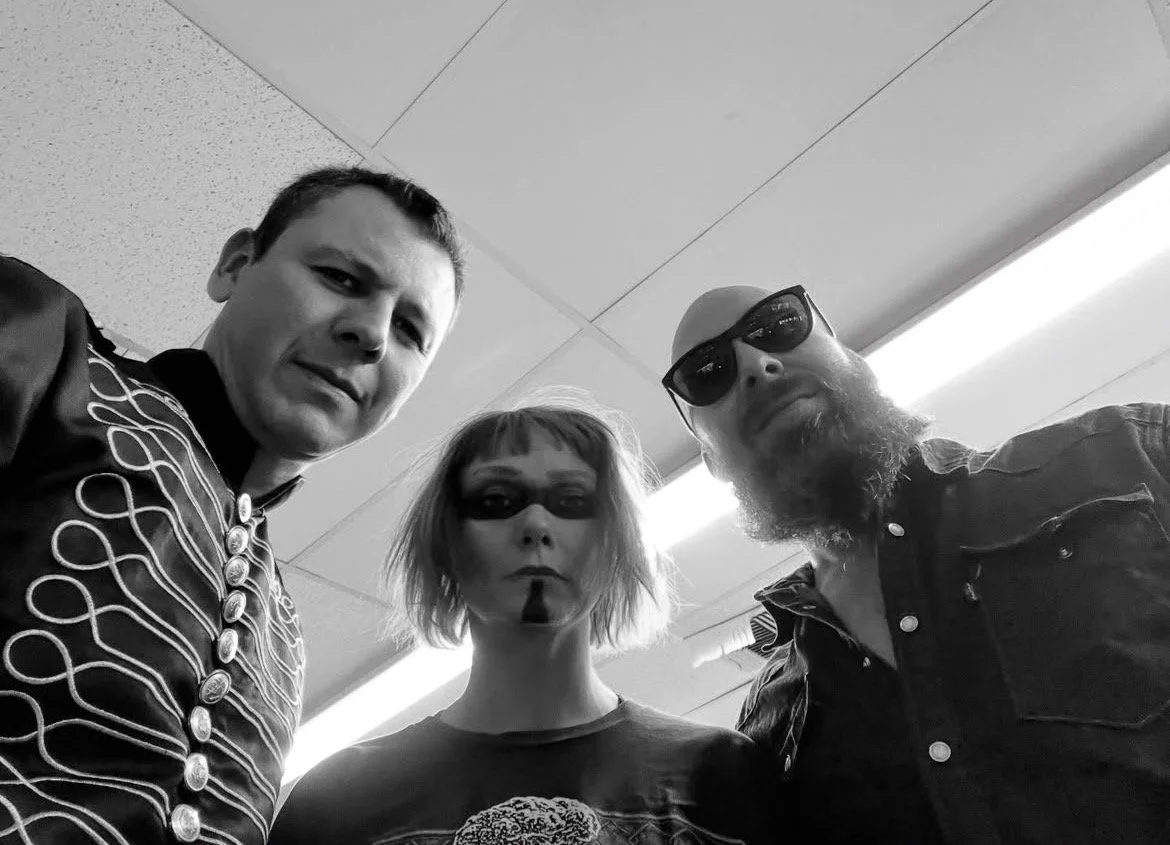 Black and white photo of three people looking down at the camera, two men and one woman, in an indoor setting. My Mistress at Rio Market Chace Levi Lacey Kay Levi Martin leal