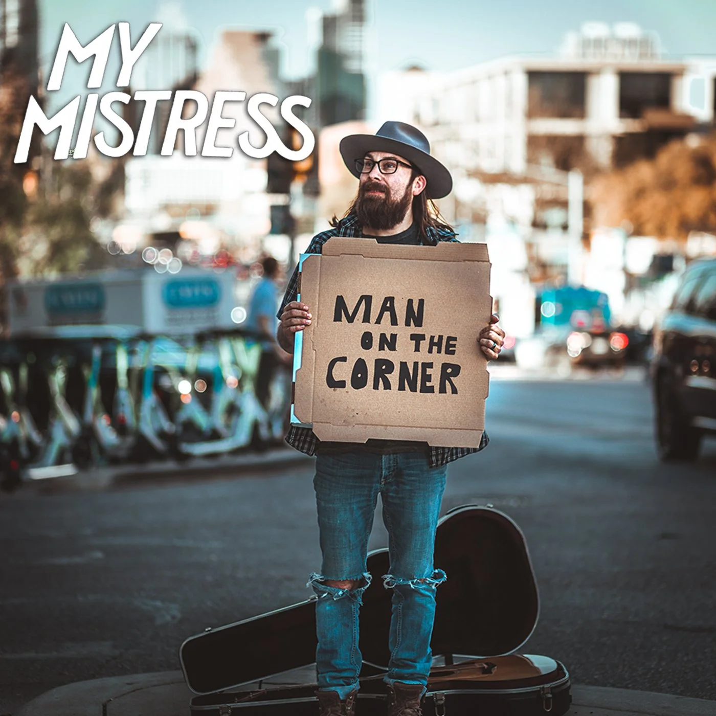 A man with long hair, beard, and glasses wearing a hat and jeans, holding a cardboard sign that reads 'Man on the corner' on a city street. Mano on the corner album cover. Chace Levi Lacey Kay Levi Geoff Garcia