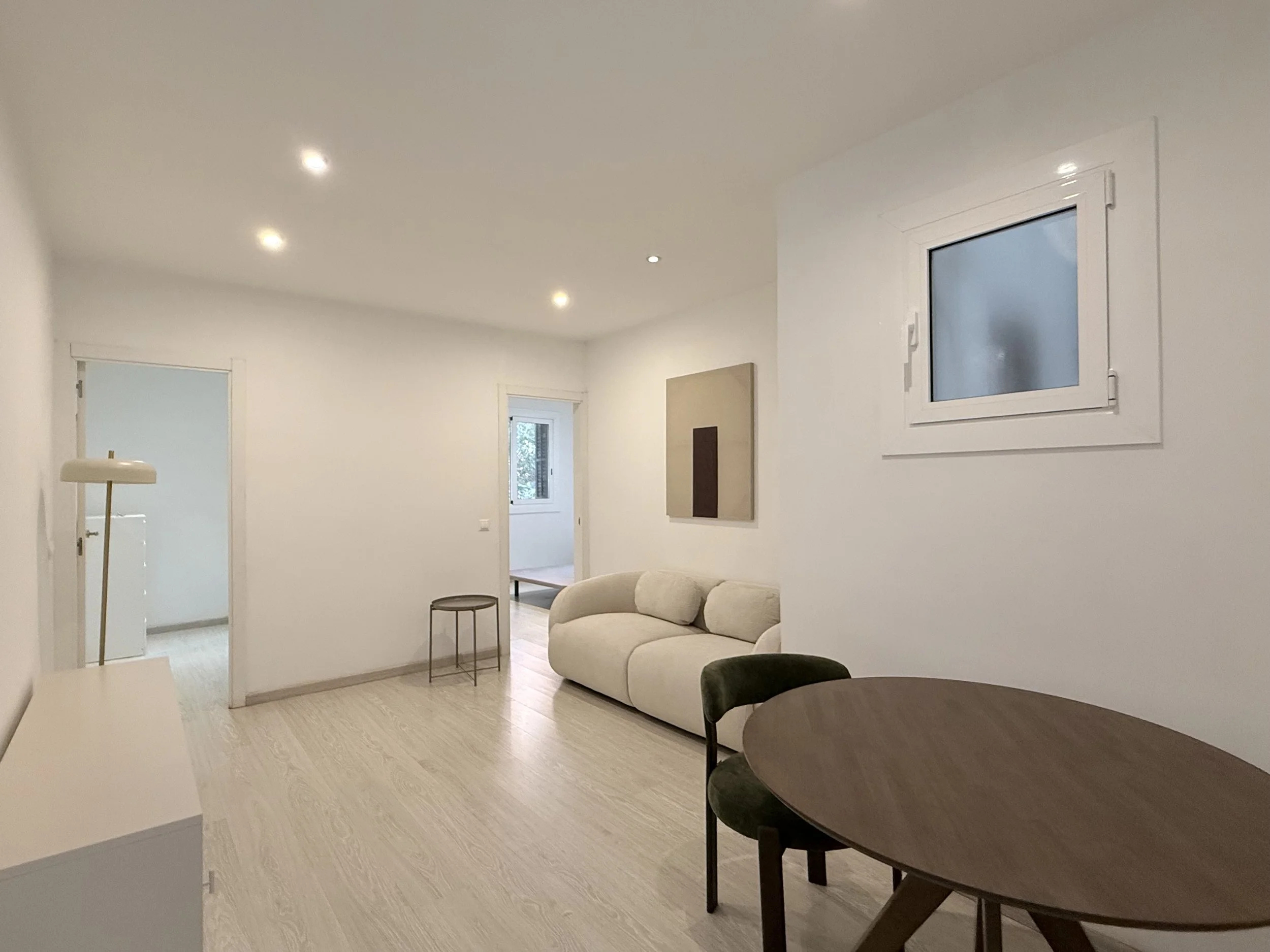 Sala de estar con sofá gris, mesa blanca y vista de balcón con sillas, paredes en tonos neutros, estantería y ventana grande con cortinas.