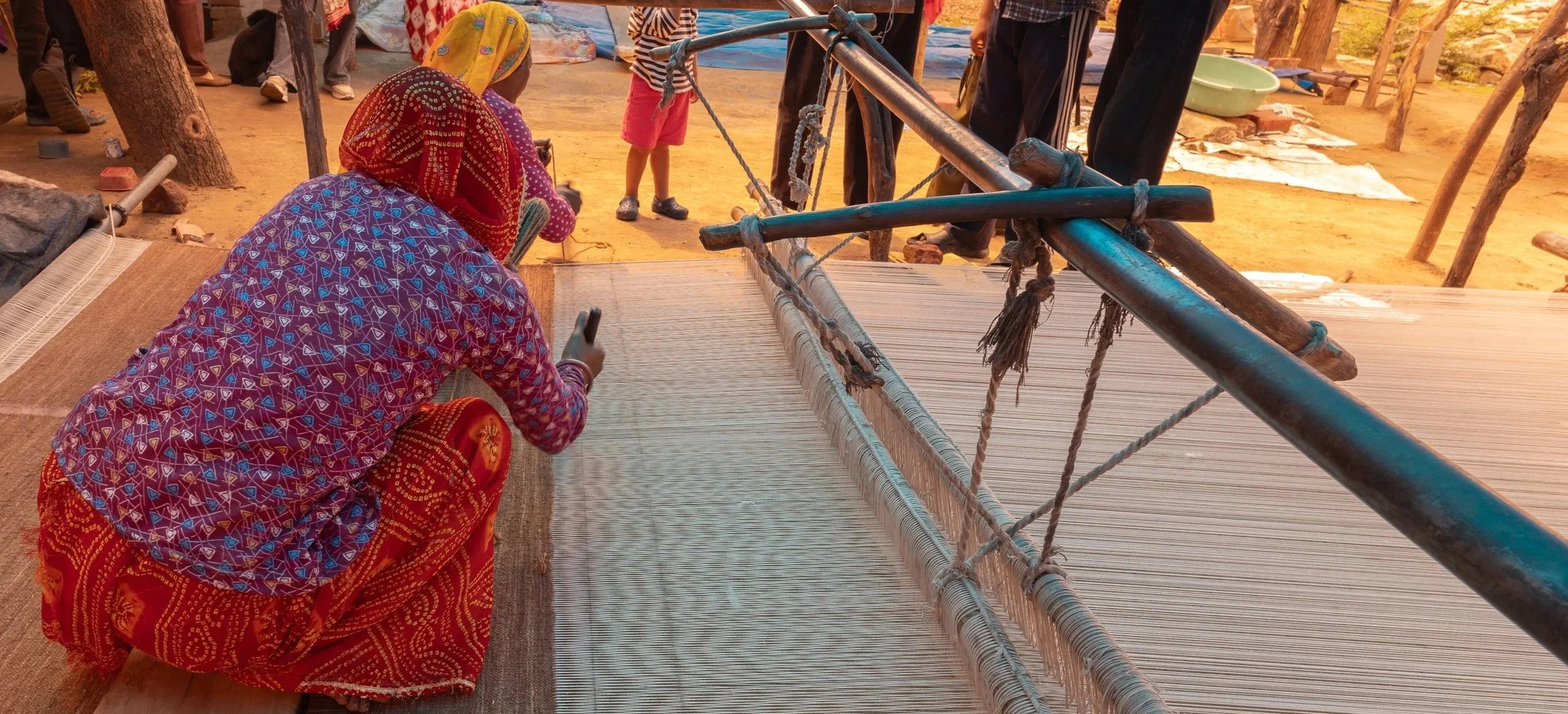 A woman wearing traditional colorful clothing is sitting on the ground, using a smartphone, near a large loom with a woven fabric. Several people are standing and walking around on a dirt ground, with some colorful fabrics and objects hanging or laid out around the area. The setting appears to be outdoors, possibly at a market or craft site.