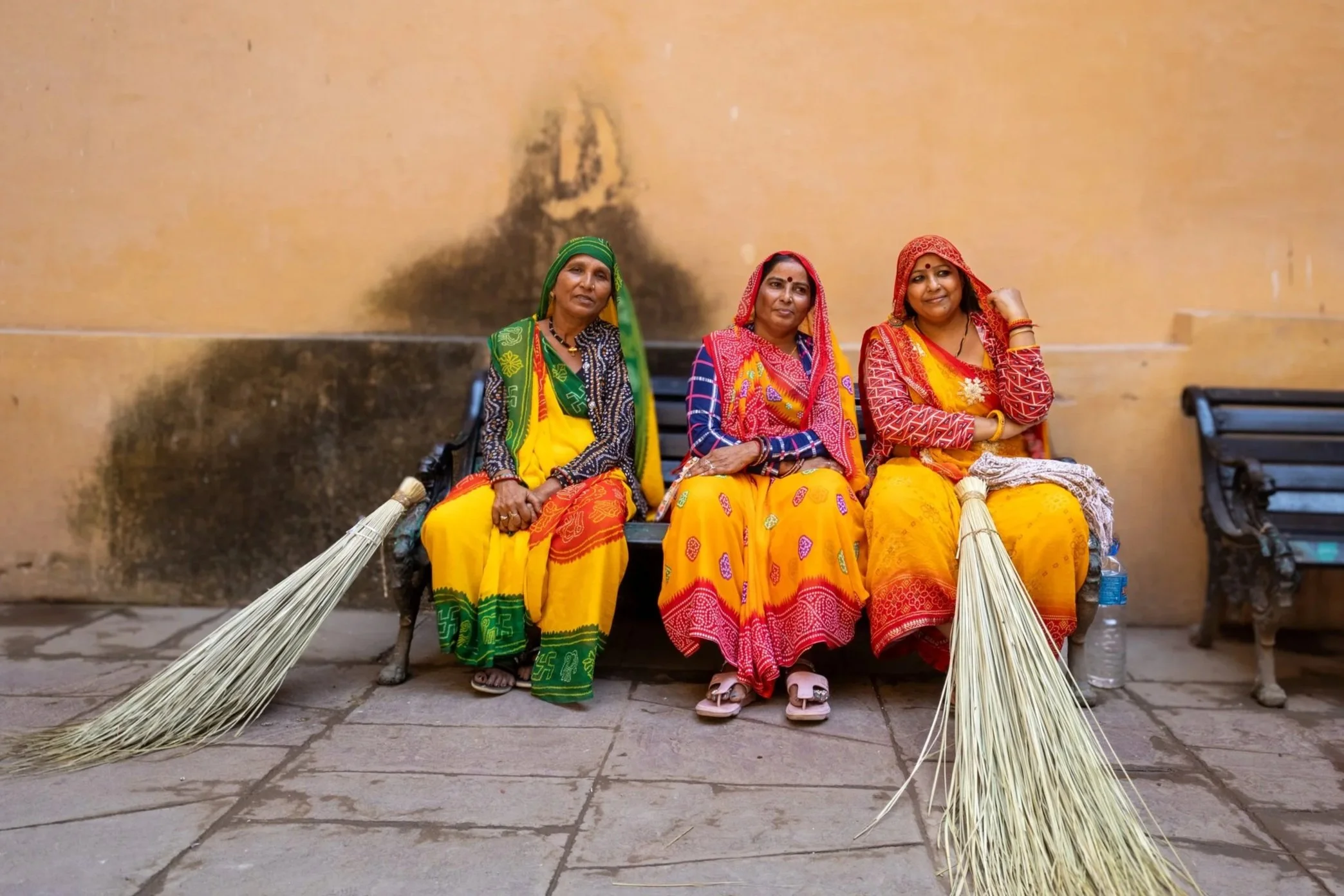 Three women in traditional colorful Indian attire sitting on a public bench against a beige wall, with two brooms placed on the ground beside them.