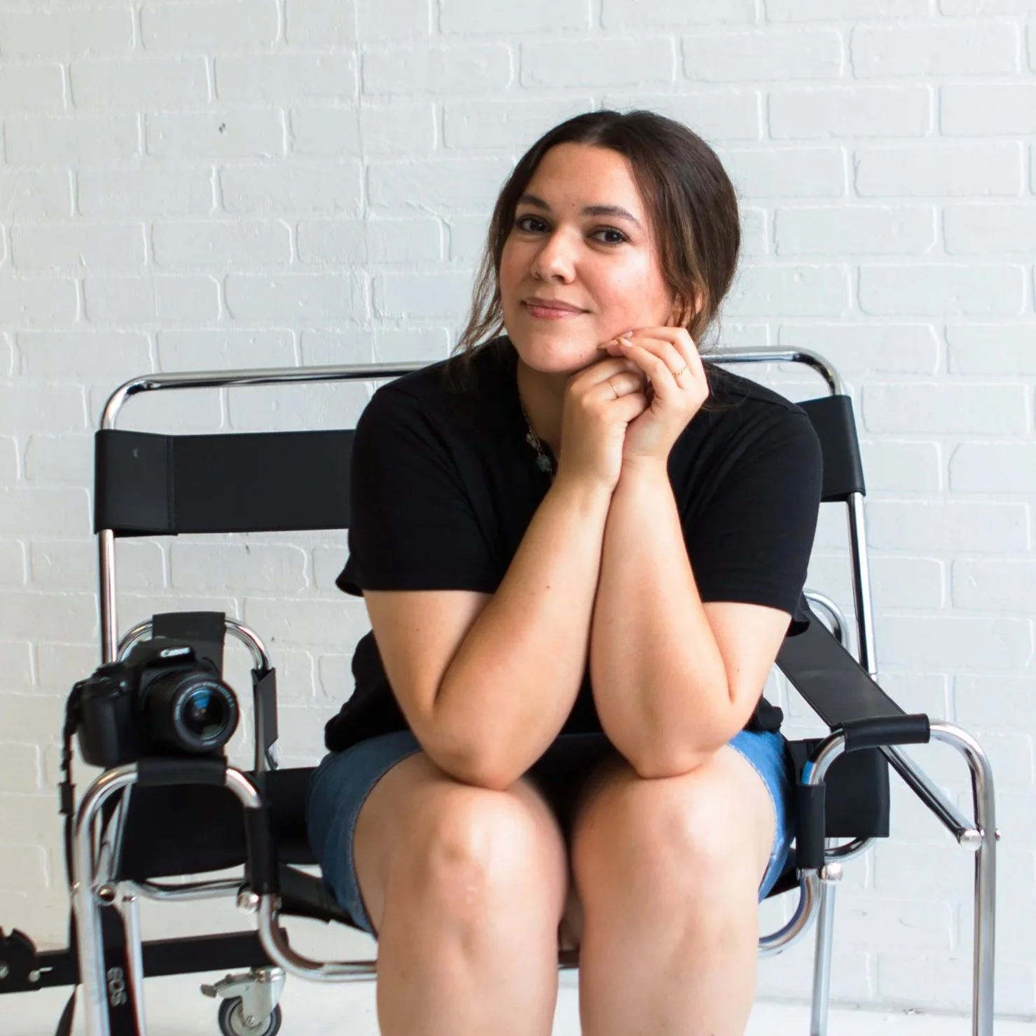 A woman sitting on a black folding chair in a photography studio, with a camera placed on the chair next to her and studio lighting equipment nearby, against a white brick wall.