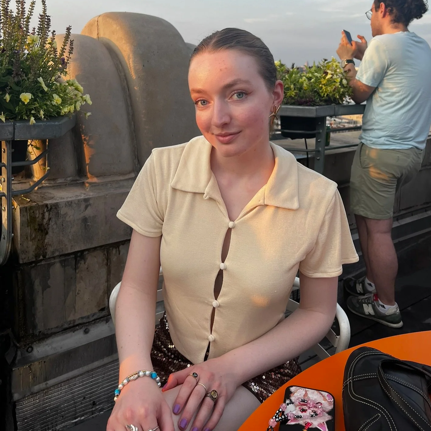 A young woman with blue eyes and fair skin, wearing a cream-colored short-sleeve blouse with buttons, sitting at an orange table with a black purse and a phone with a floral case, on a rooftop during sunset. A man with long dark hair, glasses, light blue shirt, and green shorts, is in the background taking a photo with a smartphone.