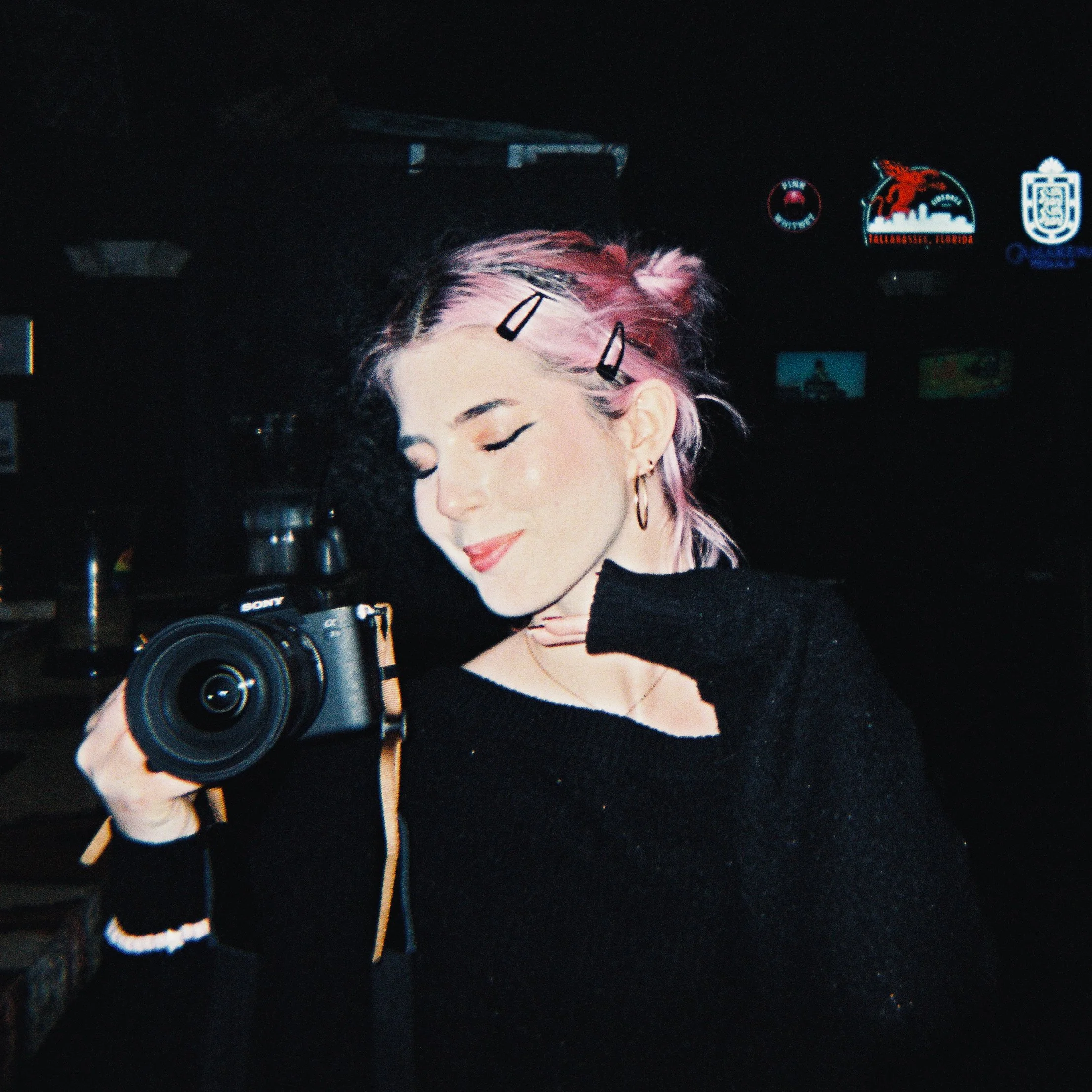 A young woman with short blonde hair and hoop earrings smiling at the camera, wearing a black sweatshirt, in a dimly lit indoor setting.