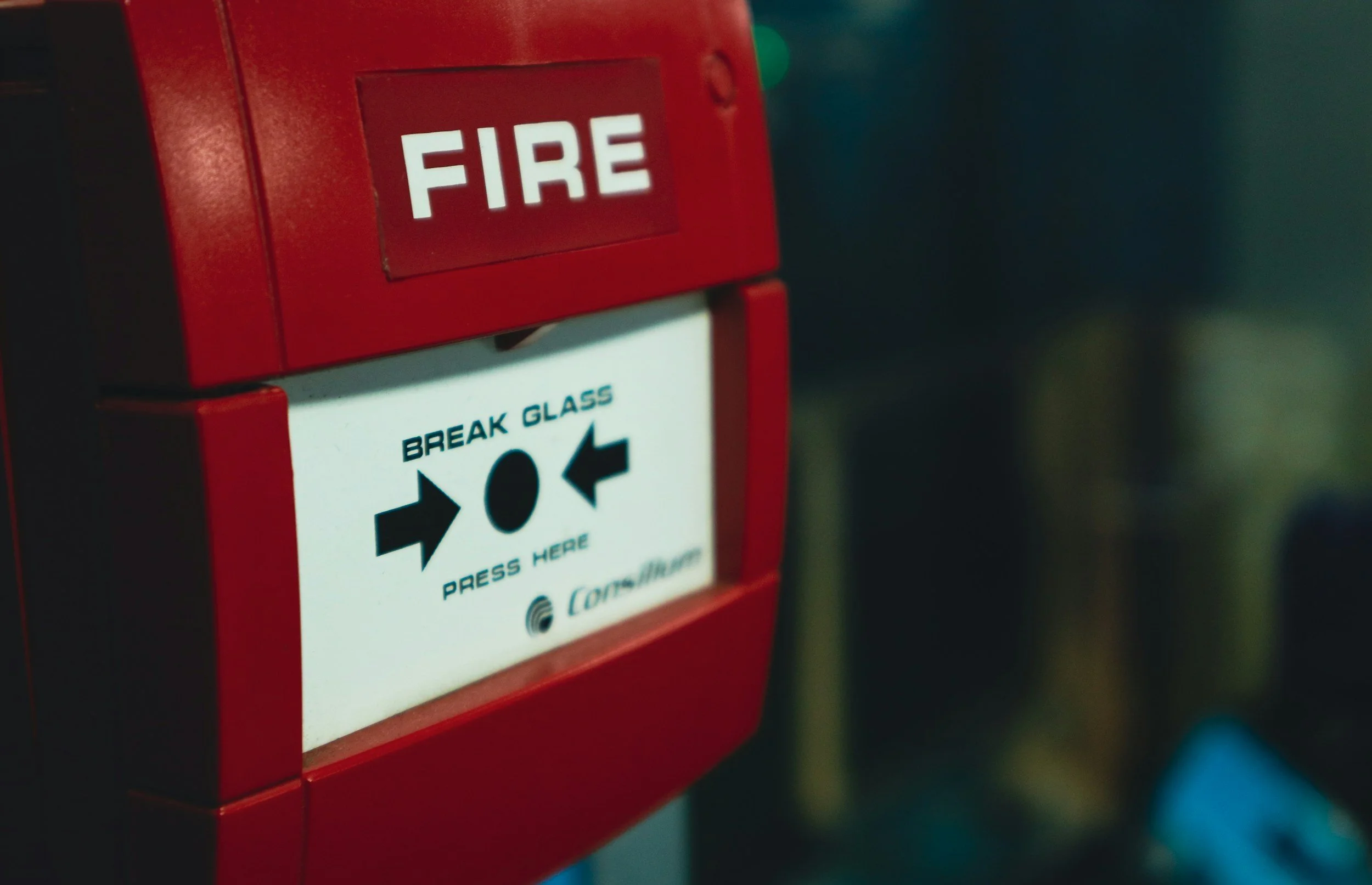 Red fire alarm with a white label that says "Break Glass" and instructions to press here, located on a wall.