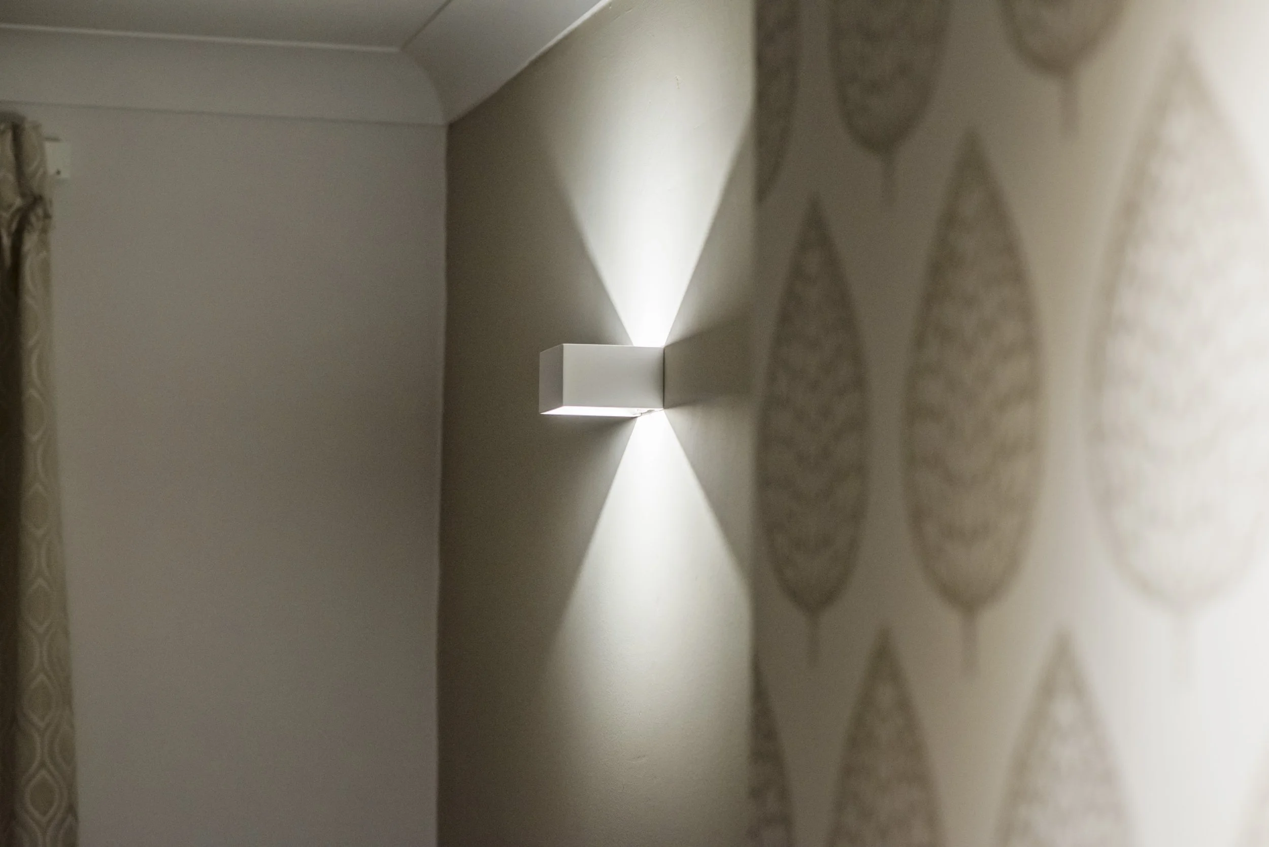 A modern wall-mounted light fixture casting a triangle-shaped pattern of light and shadow on a wall with patterned wallpaper.