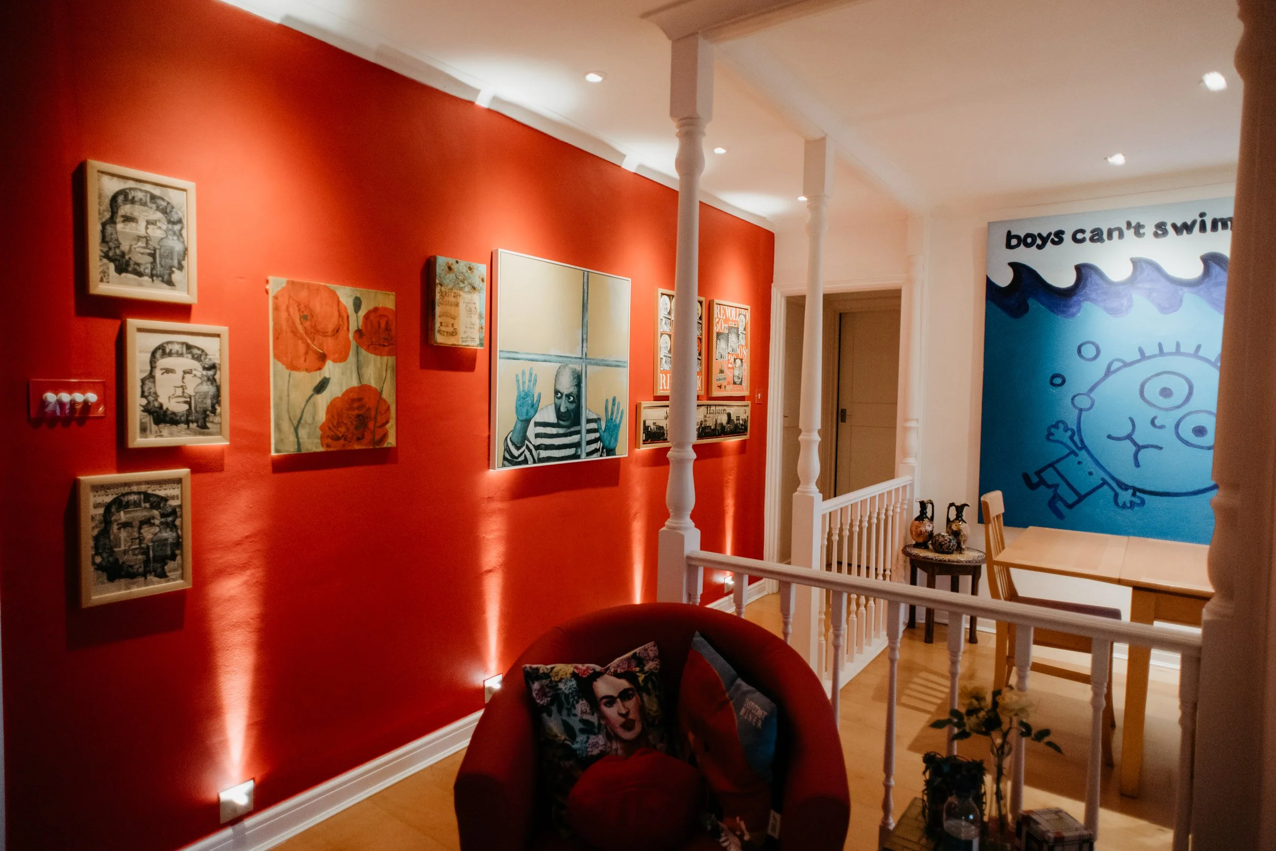 Living room with red accent wall displaying various artworks, including a mix of paintings and photographs, and a seating area with a decorative pillow featuring a portrait. There is a blue wall with cartoon-like drawings and text that says 'boys can't swim,' and a dining table with chairs in the background.