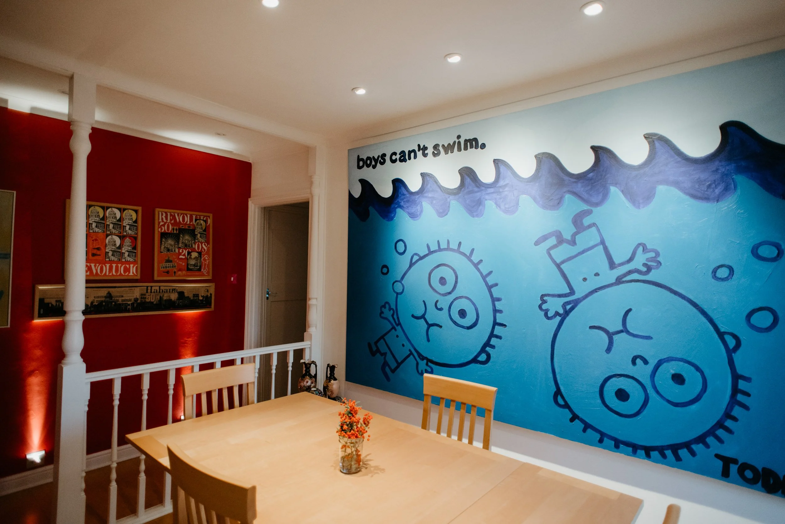 A brightly lit room with a wooden dining table and chairs, a colorful cartoon-style mural on the wall depicting a happy boy swimming underwater with a tail, and the phrase "boys can't swim." in black letters. On the red wall nearby, there are framed 