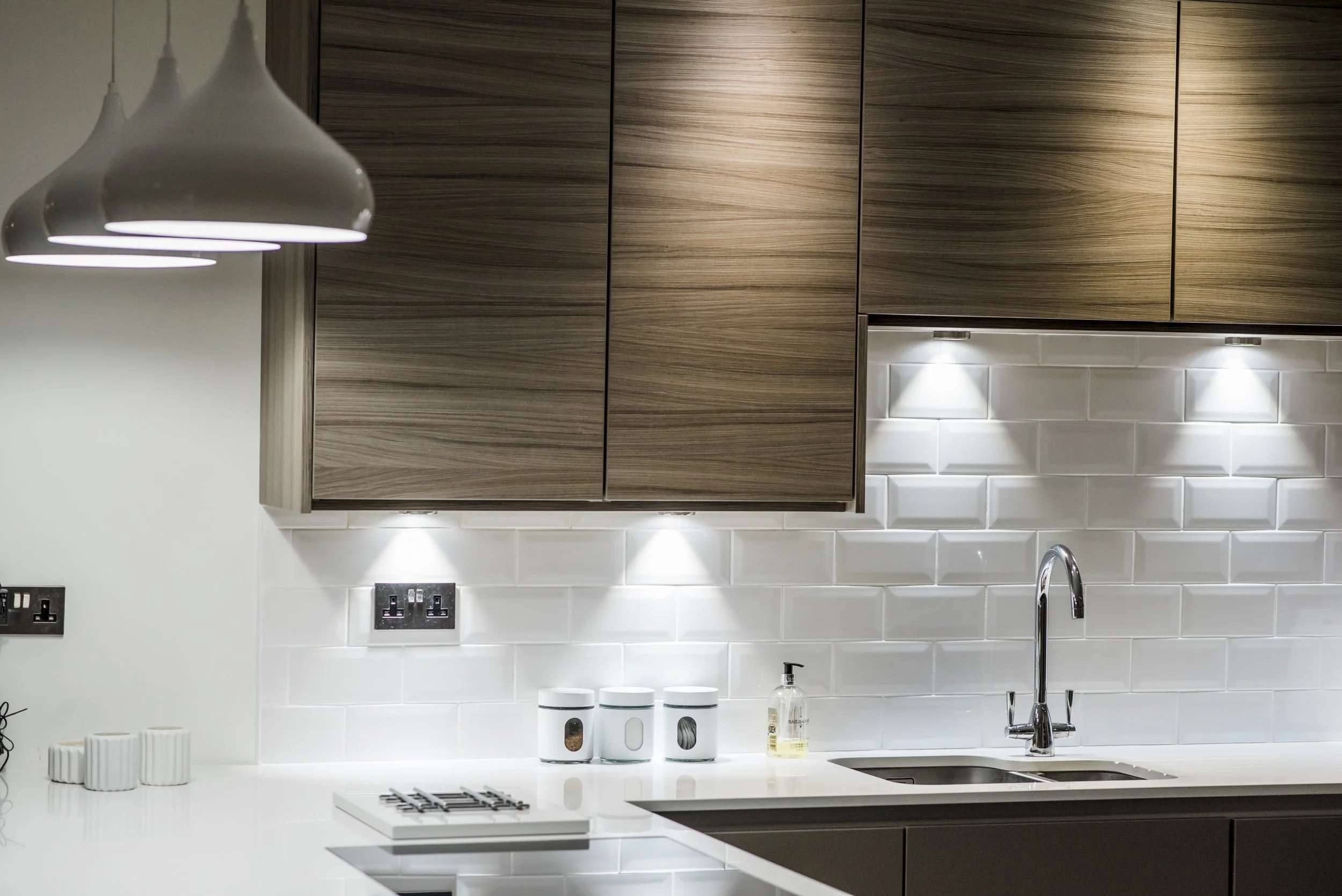 Modern kitchen with white subway tile backsplash, wooden upper cabinets, white countertop, double sink, chrome faucet, three white canisters, a soap dispenser, and pendant lights.