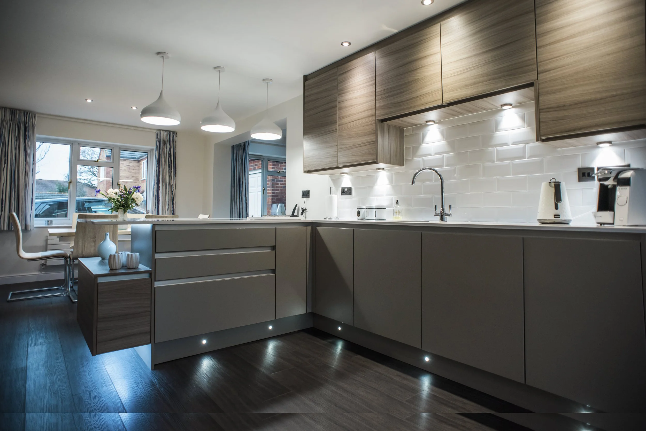 Modern kitchen with wooden cabinets, white subway tile backsplash, and under-cabinet lighting. Contains a sink, electric kettle, and coffee machine. Dining area with floral centerpiece on the table and large windows with curtains.