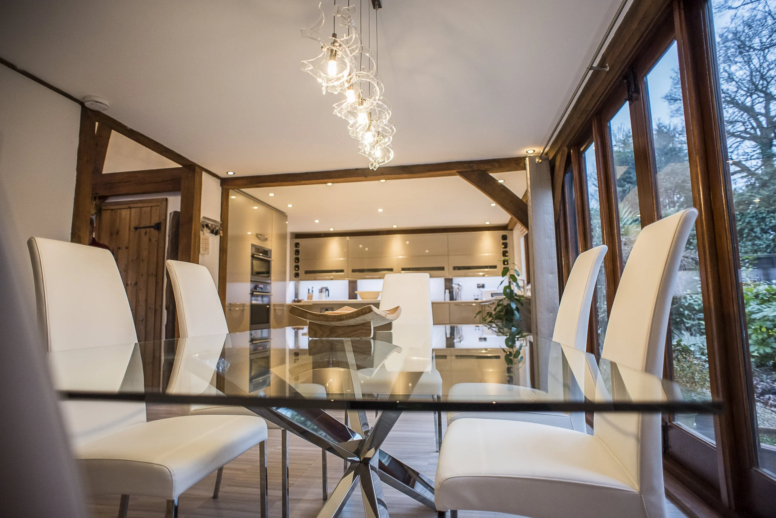 Modern dining room with white chairs around glass-top table, large windows on right side, kitchen in background, and artistic chandelier hanging from ceiling.