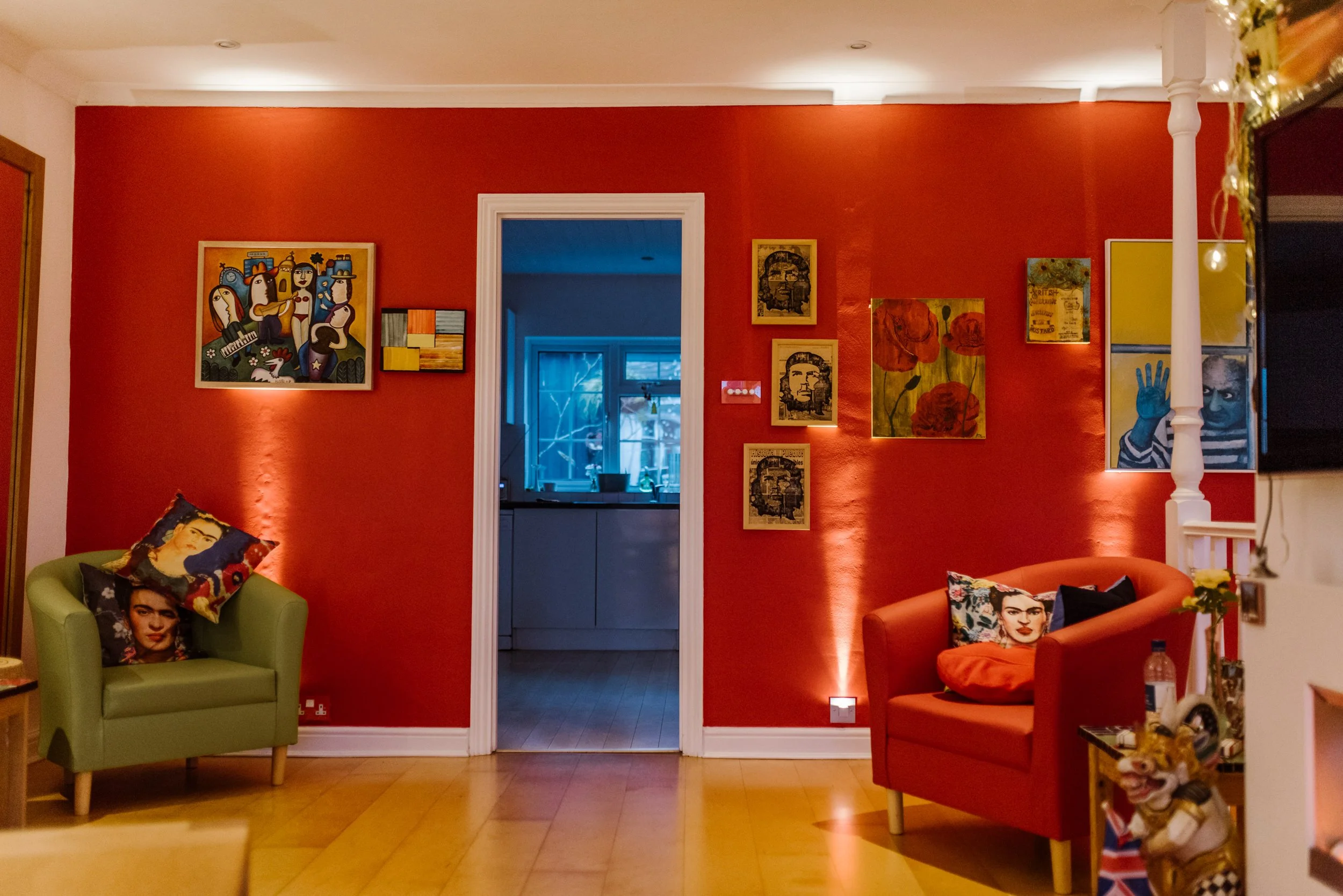 Living room with a red accent wall, framed artwork, a green armchair with Frida Kahlo pillows, and an orange armchair with a Frida Kahlo pillow. A doorway leads to a kitchen with blue lighting, and a television is mounted on the right side.