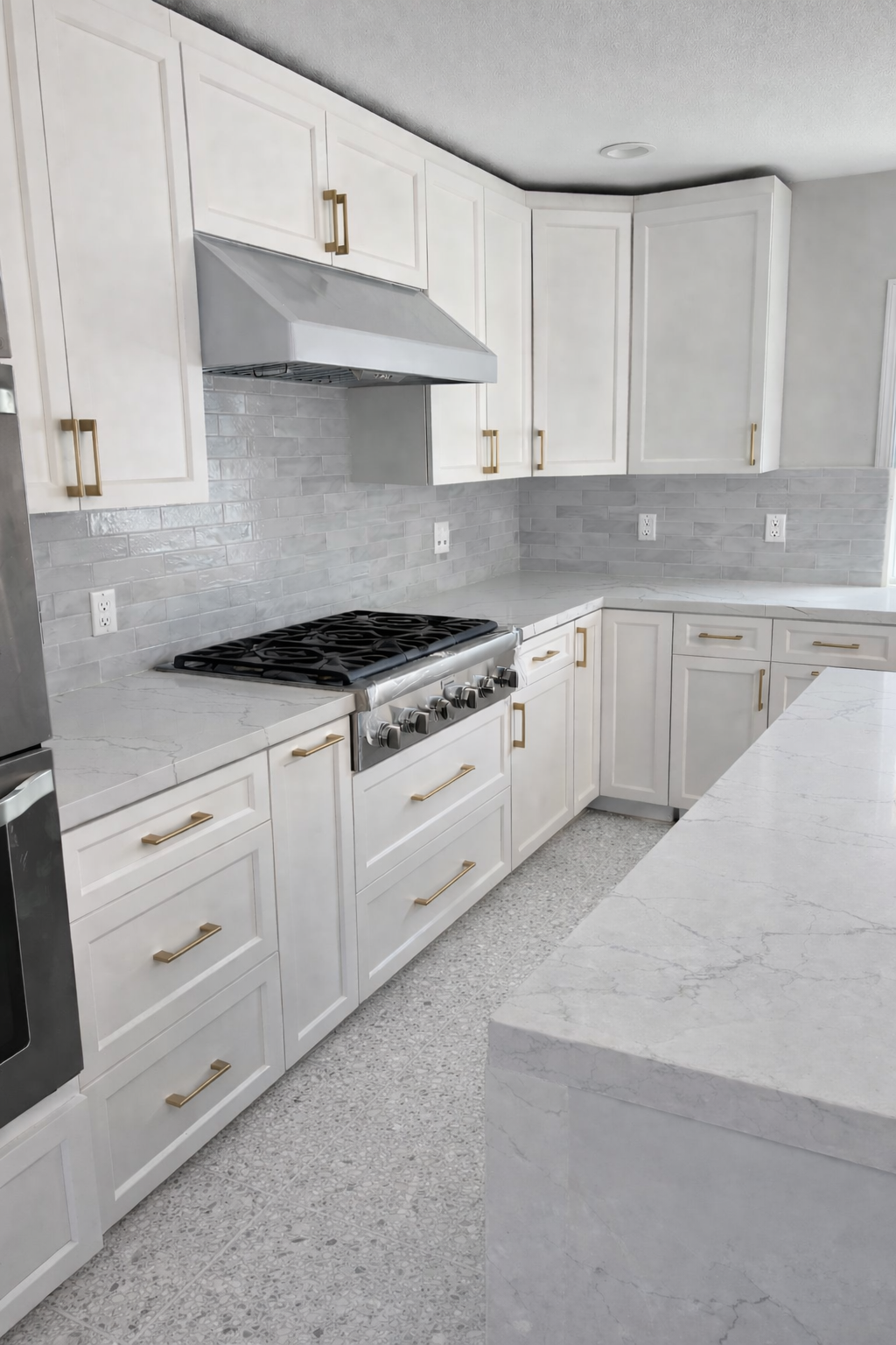 A modern kitchen with white cabinets, gray backsplash tiles, white marble countertops, stainless steel appliances including a stove and oven, and gold handles on the cabinets.