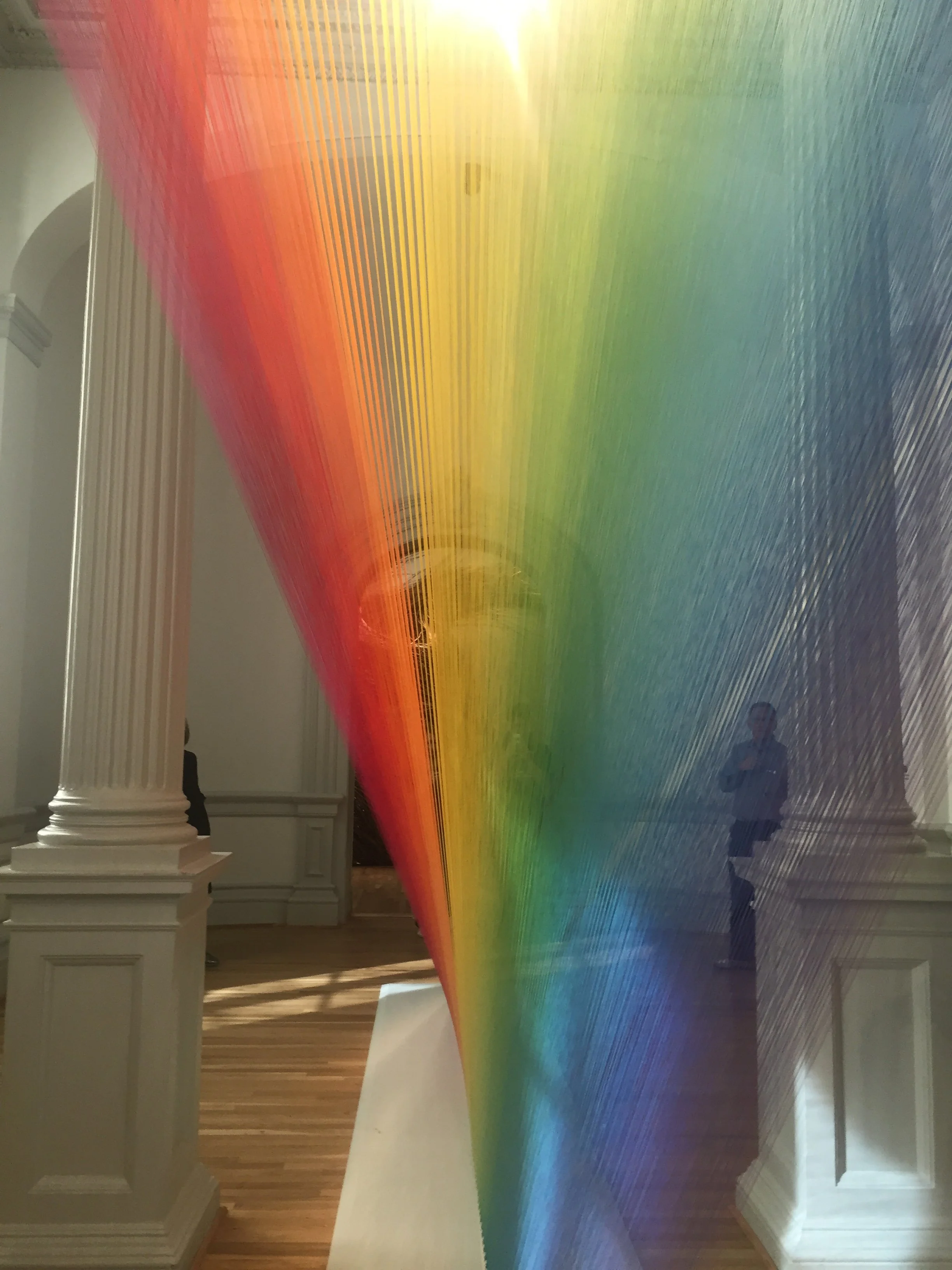 A colorful art installation featuring curved, intertwined ribbons of fabric or material in rainbow colors, creating a gradient effect on a reflective surface, with people and architectural details visible in the background.