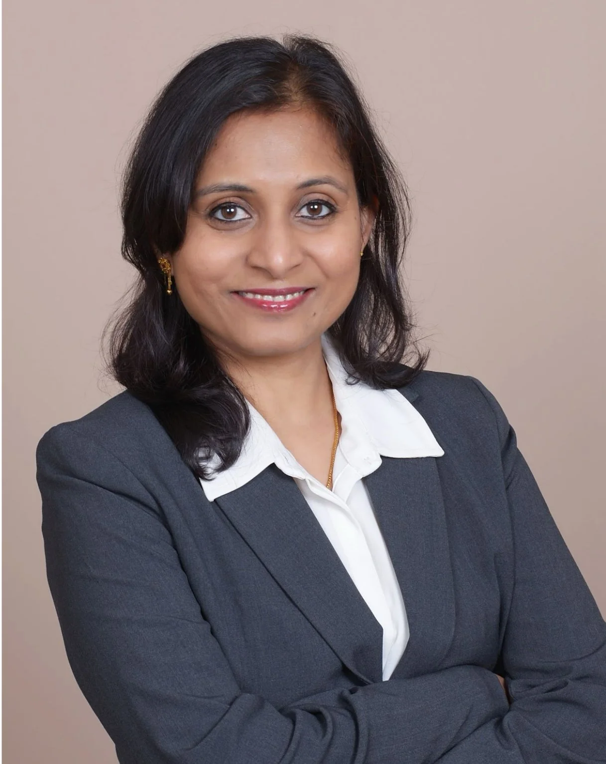 A woman in a dark blazer and white shirt, with shoulder-length dark hair, smiling with arms crossed, neutral background.