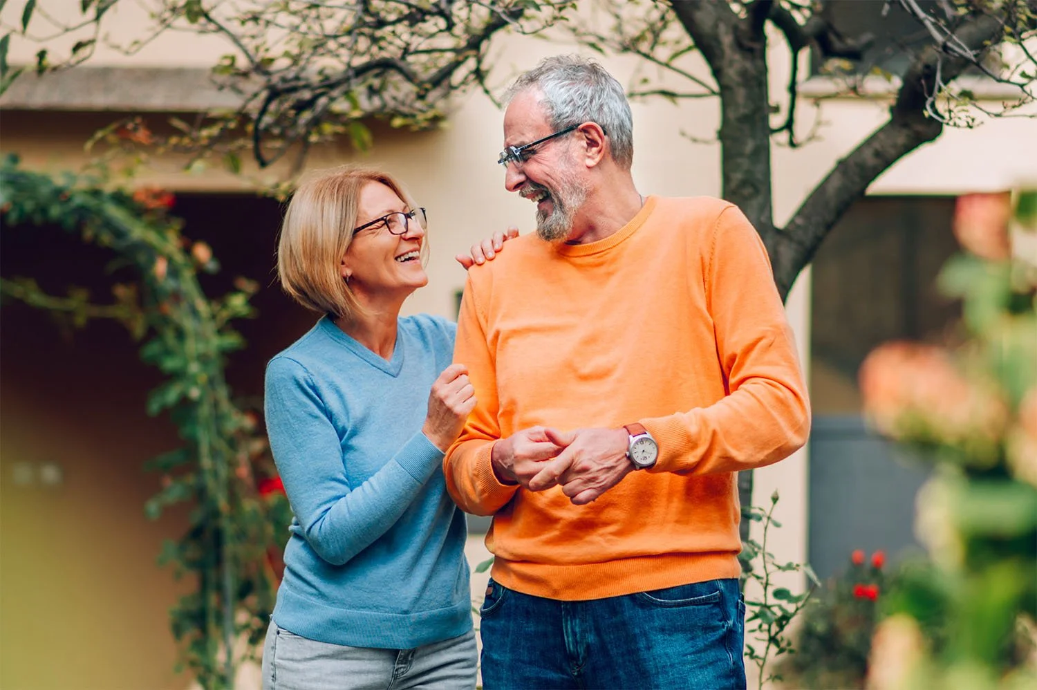 An older woman and man smiling at each other, with the woman touching the man's shoulder, in their Toowoomba garden setting with trees and flowers. The woman has short blonde hair, glasses, and is wearing a blue sweater.