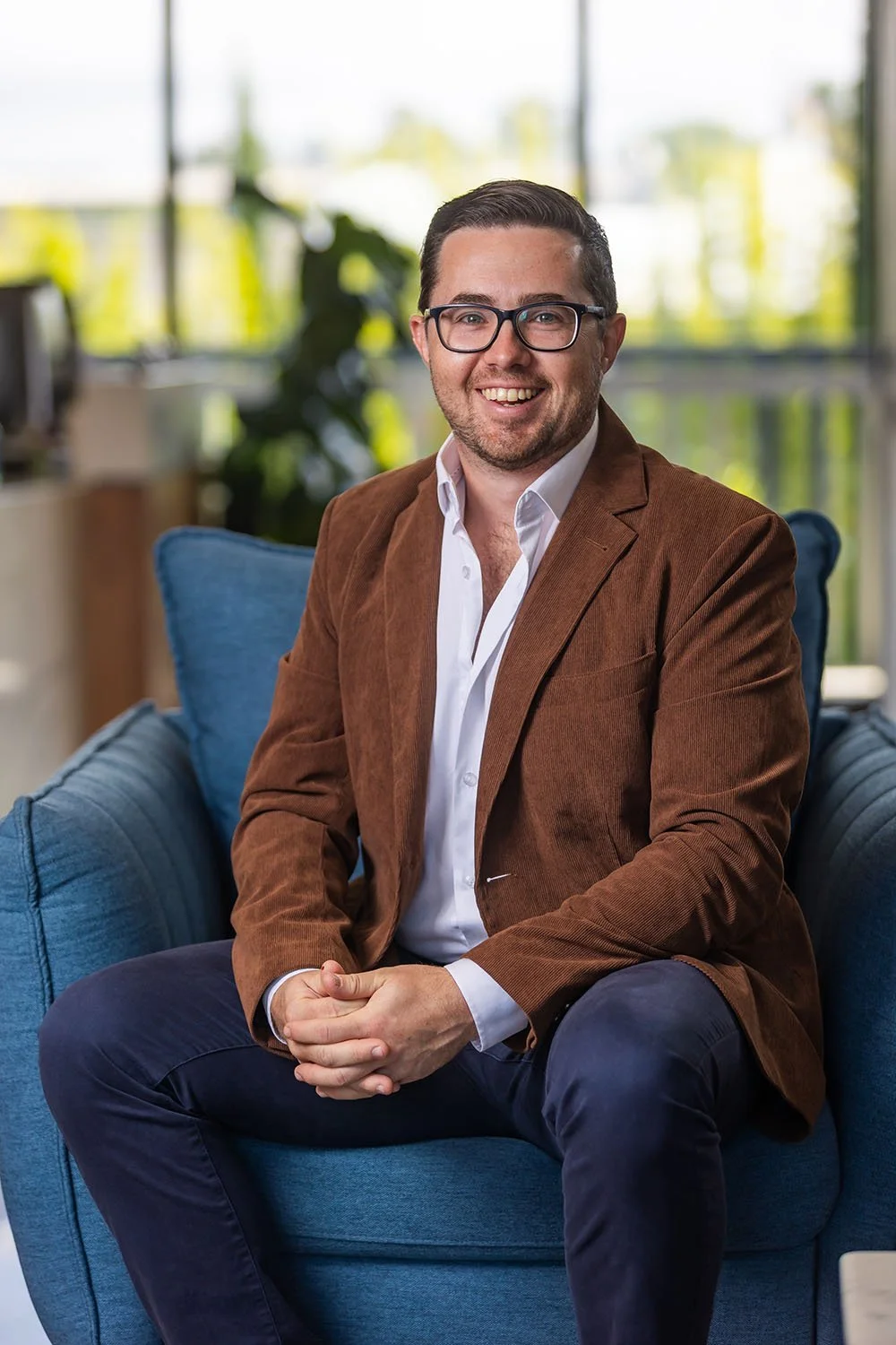 Financial Planner Peter Walters in a dark suit, light blue shirt, and glasses sitting on a blue couch, smiling at the camera, with a window showing an outdoor view in the background.