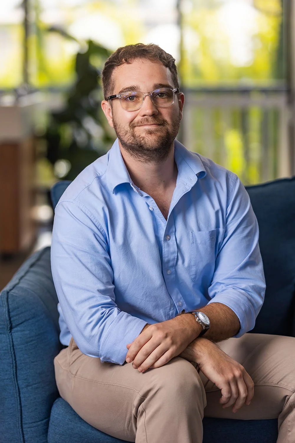Client Service Officer Jarrad Mason smiling with glasses sits on blue sofa in a well-lit room with large window, wearing a light blue shirt, beige pants, and dark shoes.