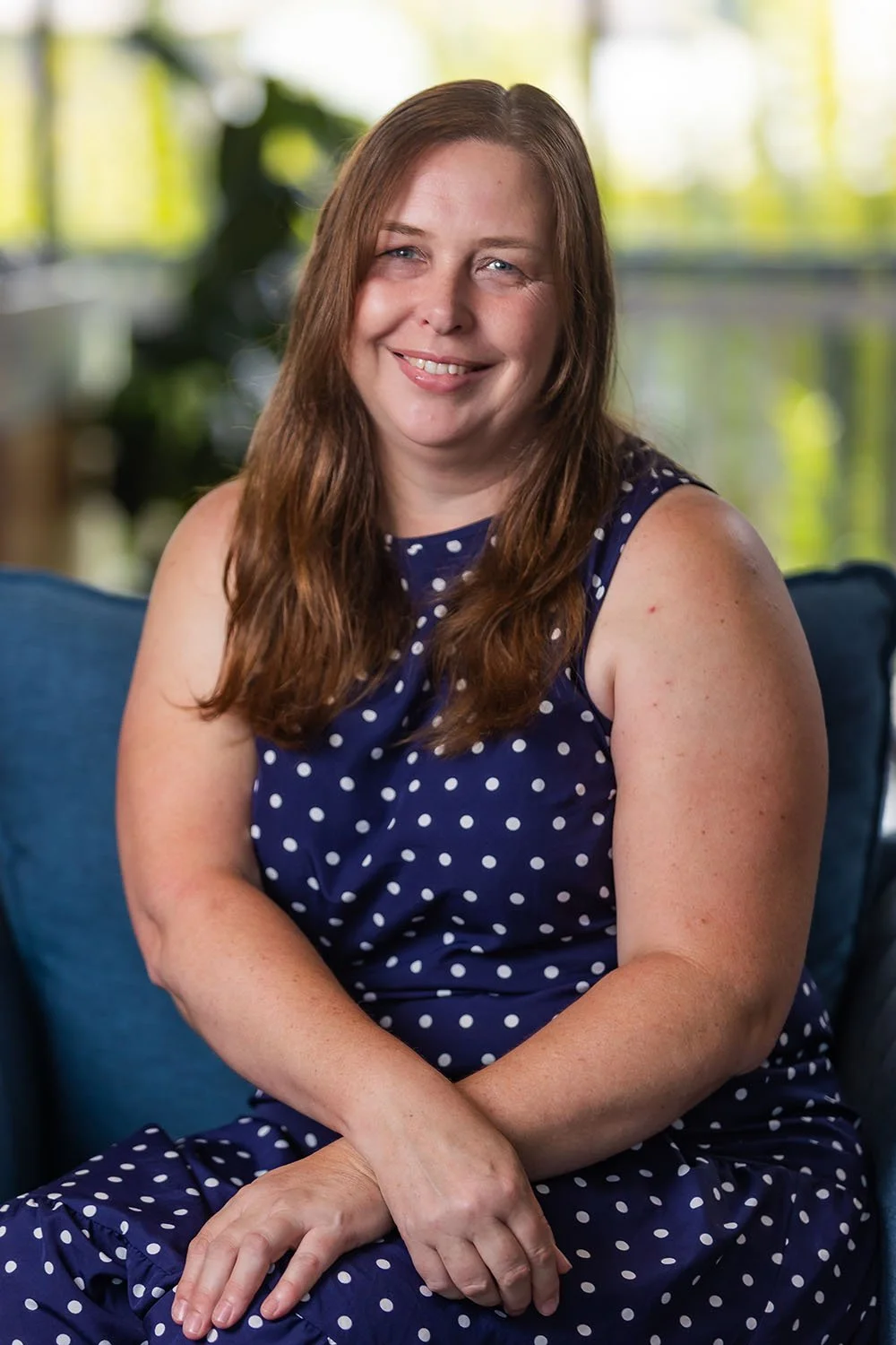 Client Service Officer Kristy Churchill with long brown hair, smiling, sitting comfortably on a blue sofa in a well-lit room, wearing a bright blue blouse and beige pants.