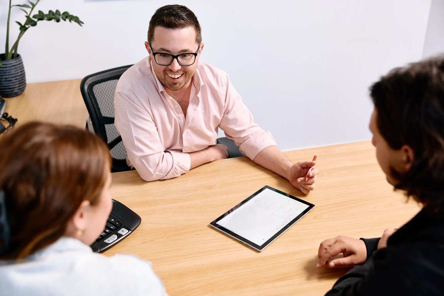 Financial Adviser Pete Walters smiling and holding a stylus, having a meeting with clients. The two clients are facing Pete, sitting at a wooden table with a tablet and a phone.