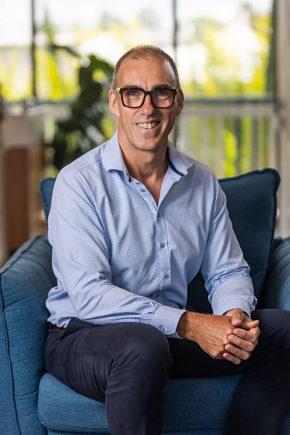 Financial Planner Stan Moffat with glasses, smiling, sitting on a blue armchair in an office with large windows and a city view.