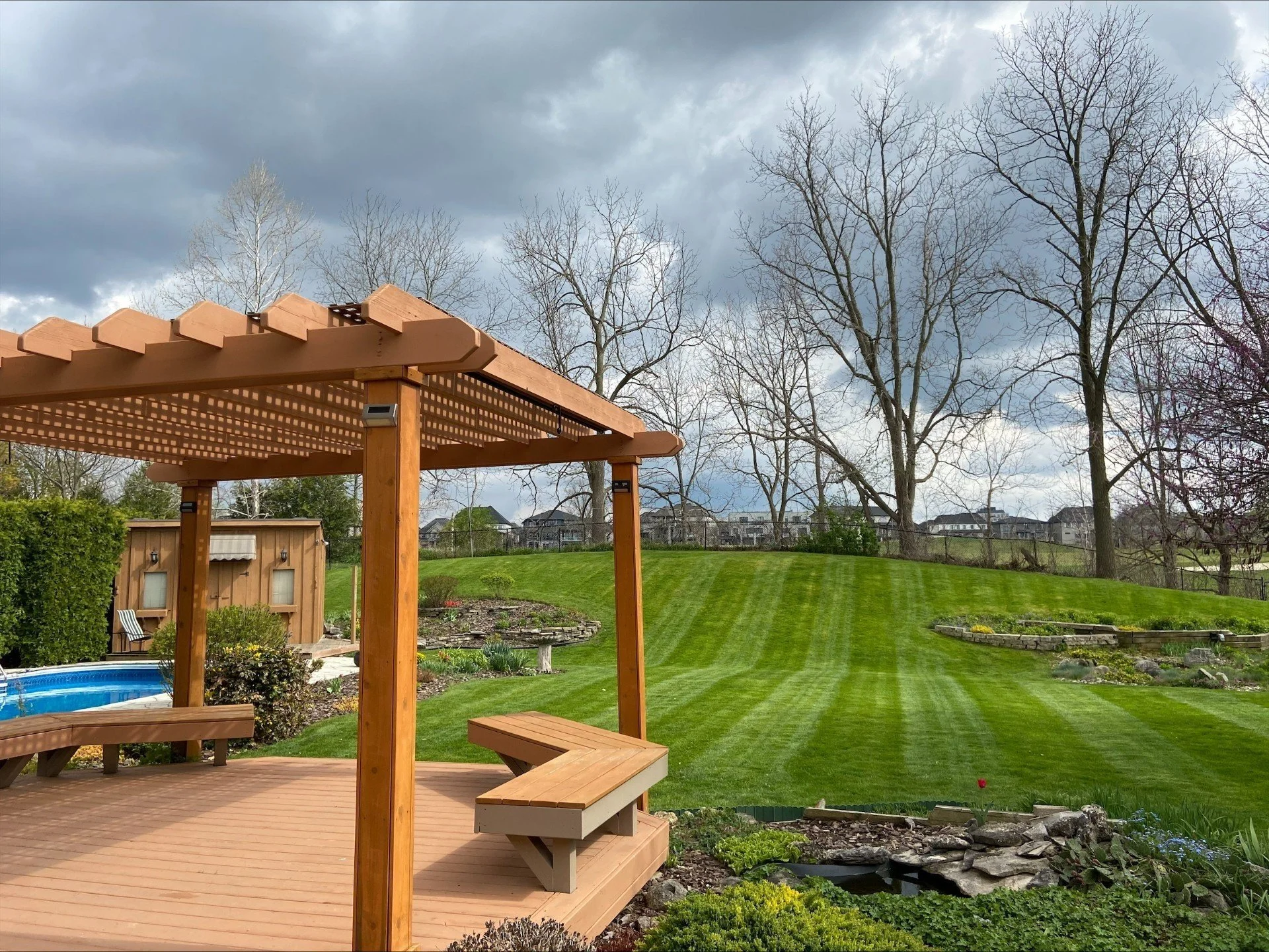 A backyard with a wooden pergola on a deck, green grass, a small pond, a storage shed, and leafless trees under a cloudy sky.