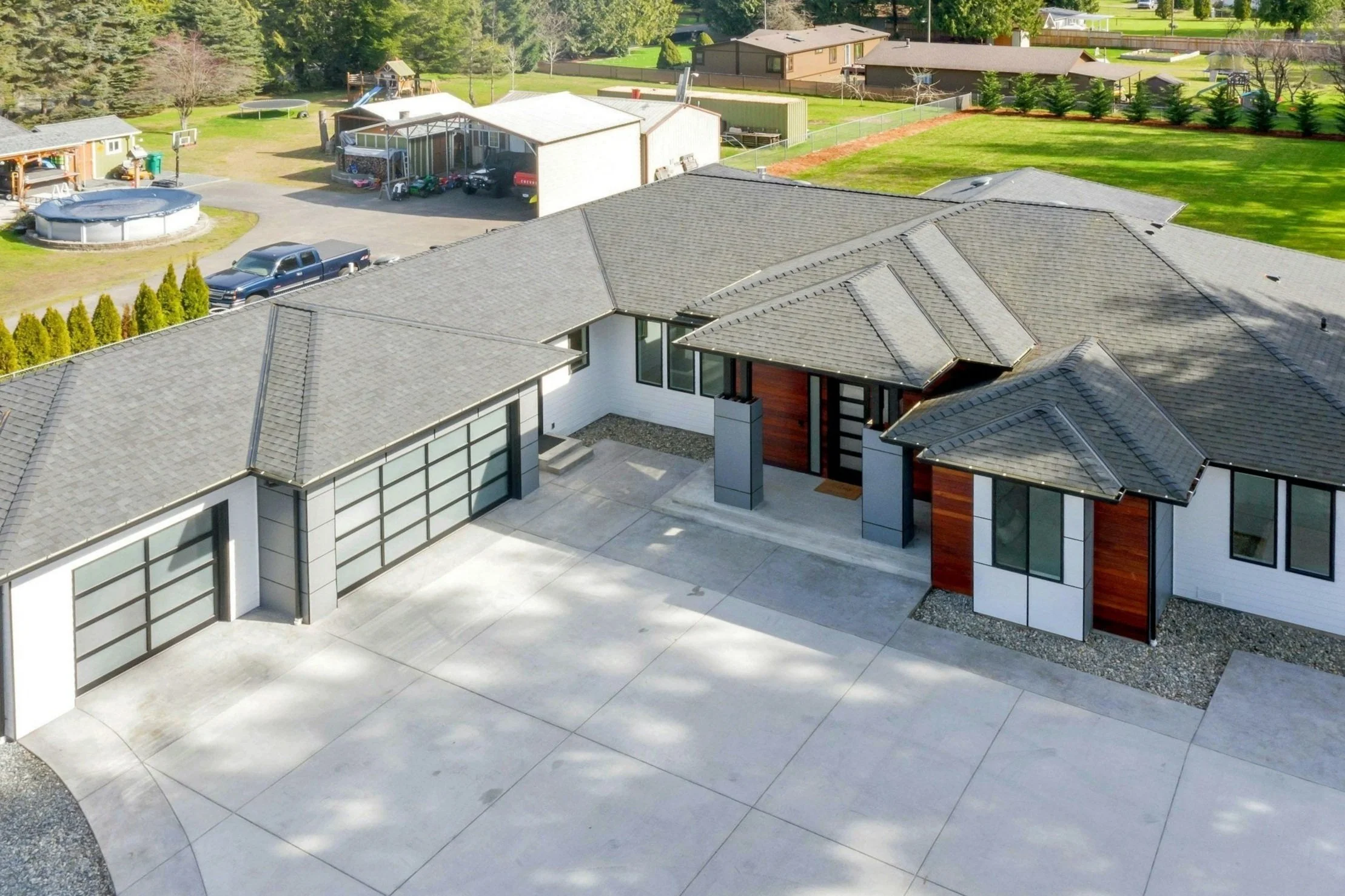 Modern house with a gray tiled roof, white and wood exterior walls, large front driveway, and spacious backyard with greenery.
